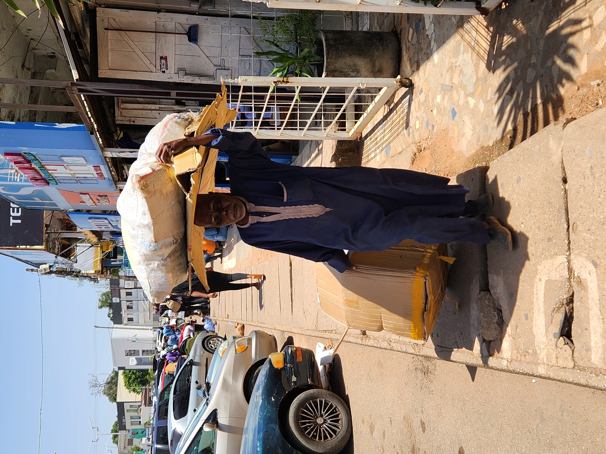 An elderly Black man wearing a dark blue robe is carrying a large sack on his head, balanced on a piece of cardboard, and holding a cardboard box in his right arm. He is standing on a concrete sidewalk next to a busy street with parked cars. The street is lined with buildings, some with storefronts, and cars driving on the street. The scene is in the city of Jos, Nigeria.