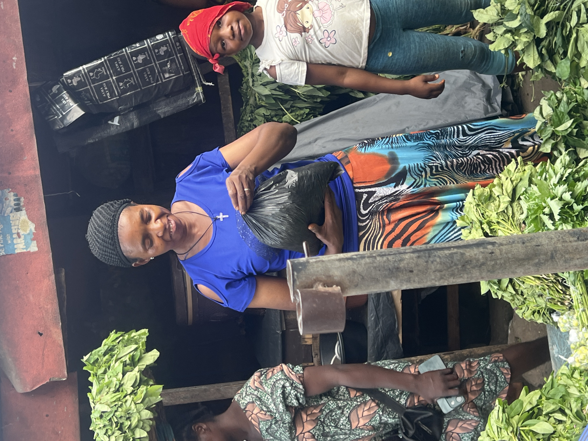 The image shows a woman in Nkpor, Nigeria, likely in a market setting. She wears a blue, cut-out shoulder shirt and a colorful, patterned skirt. A black knitted cap covers her head. She holds a black plastic bag, possibly containing purchased goods. Her expression is friendly and she appears to be interacting with someone.
A young girl in a white shirt with a cartoon graphic and blue pants stands near the woman, wearing a red headscarf. Another person in a patterned dress is partially visible at the bottom of the frame, holding a mobile phone.
Bundles of leafy green vegetables are visible, suggesting the location is a marketplace. A black plastic bag with white writing on it hangs in the background. The surrounding environment includes a metal or wooden support structure, suggesting a stall or booth.