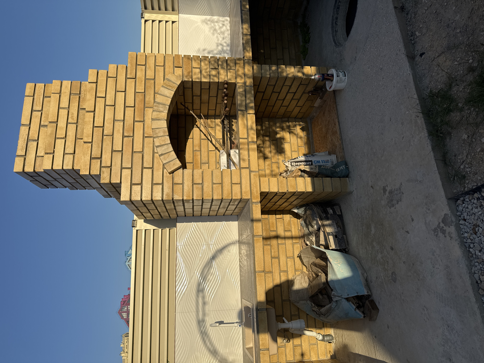 The image shows an outdoor brick barbecue grill with a sink area. The grill is constructed from yellow bricks and features an arched opening for the fire. Above the opening, there is a chimney that tapers as it rises. To the left of the grill is a countertop with a sink and faucet. Below the countertop, a bag of building material, possibly cement or sand, is visible, along with some wooden debris. The ground is concrete, with a circular opening to a pit on the right side of the image. In the background, a light-colored fence separates the area from houses with colorful roofs. The sky is clear and blue. The overall scene depicts an unfinished outdoor kitchen or barbecue area in a residential setting. The location is identified as Uyutnoye, Ukraine.