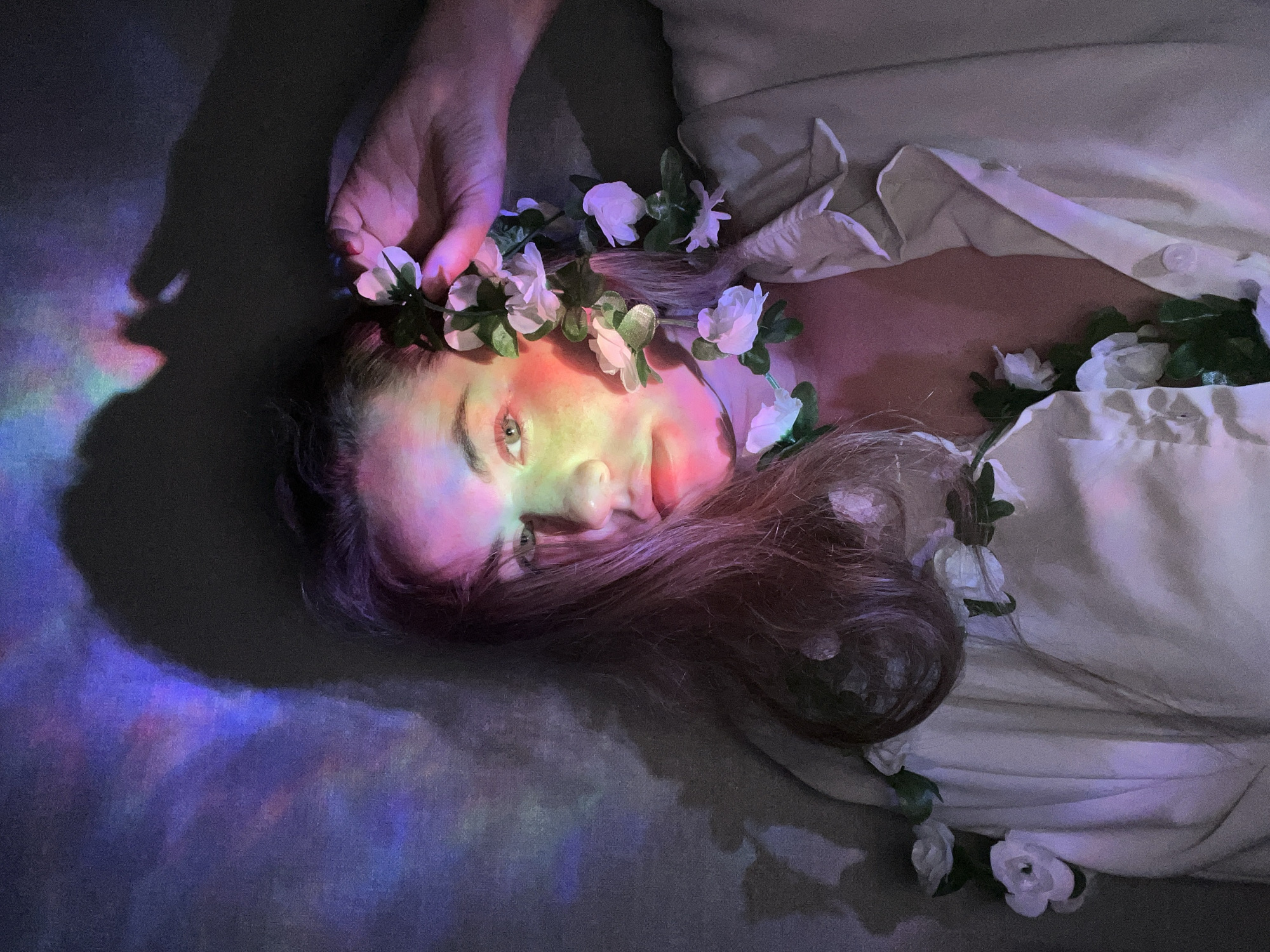 A woman with long brown hair lies on a gray surface, possibly a floor or bed, wearing a white garment resembling a shirt or dress. She is adorned with a flower crown of white roses and green leaves. A hand, presumably belonging to another person, is positioned near her head, touching the flower crown. The scene is bathed in a soft, multicolored light, creating a rainbow effect on her face and the surrounding area. The lighting suggests an indoor environment. The image does not contain information that can verify the city of Chaikovsky, Russia.