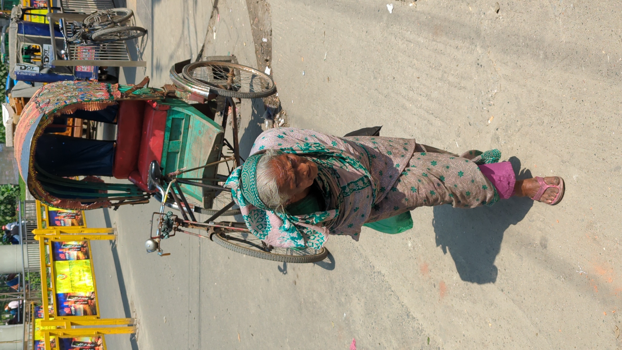 An elderly woman lies sleeping on the ground next to a rickshaw in Dhaka, Bangladesh. The rickshaw, decorated with colorful patterns, is parked on the side of a street lined with yellow barricades bearing advertisements. The woman is wrapped in a patterned shawl, wears pink pants, and rests her head on a small bag. The environment is sunny with distinct shadows.
