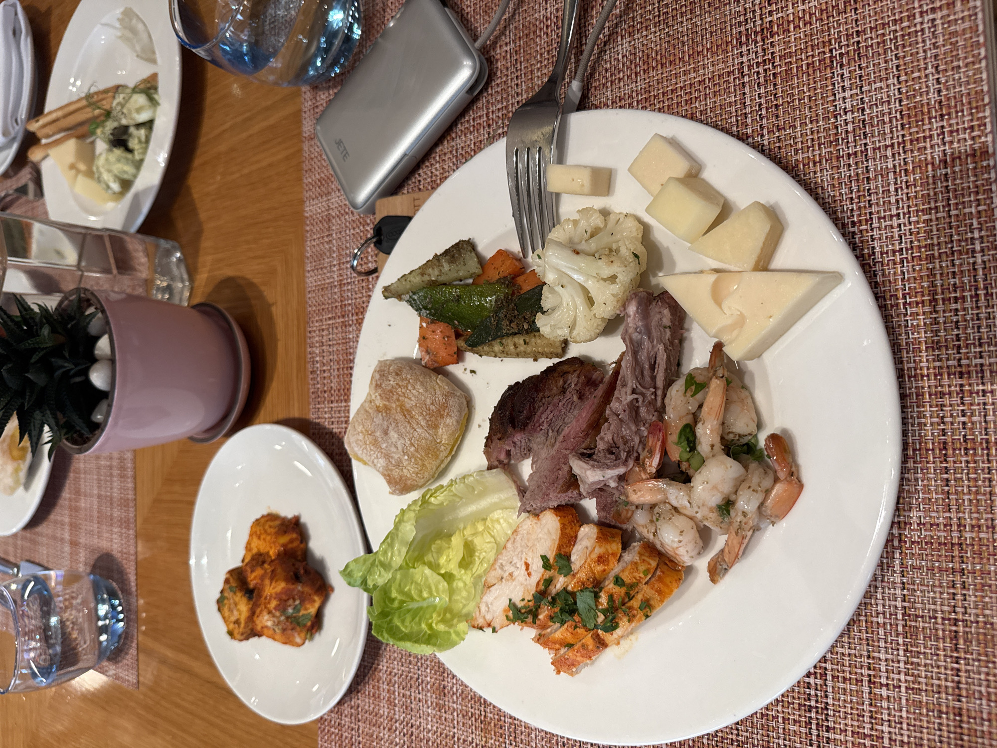 The image shows a close-up of a meal served on a white plate. The plate contains a variety of food items, including what appears to be grilled chicken or fish with herbs, sliced roast meat, cooked shrimp, and roasted vegetables such as carrots, zucchini, and cauliflower. There are also several pieces of cheese on the plate, some triangular and some cubed. A small roll or bun is also visible. To the left of the main plate, another smaller white plate holds three pieces of what looks like tandoori paneer. In the background, there are glasses of water, a small potted plant with succulents, and a portable charger. The setting appears to be a restaurant or dining area, with a wooden table and a textured placemat. The image does not contain any information that would suggest its location in Pune, India.