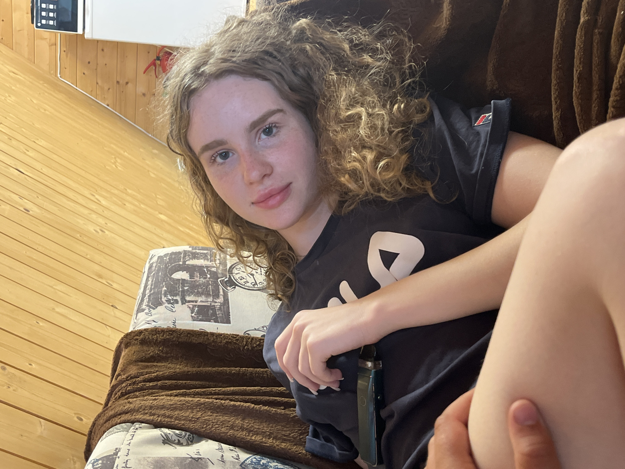 In a room with wood-paneled walls and ceiling, a young woman with curly blonde hair and blue eyes is lying on a brown sofa. She is wearing a dark t-shirt with a white logo. Her left hand is resting on her chest, holding a dark green object, possibly a remote control. A brown blanket is draped over the sofa. A person's hand is visible on the woman's leg. In the background, a white appliance with a dark top is visible, possibly a microwave, and a red fire extinguisher is mounted on the wall. The scene suggests a casual indoor setting. The image does not contain any specific landmarks or contextual clues that would indicate a location in Vasilyevo, Russia.