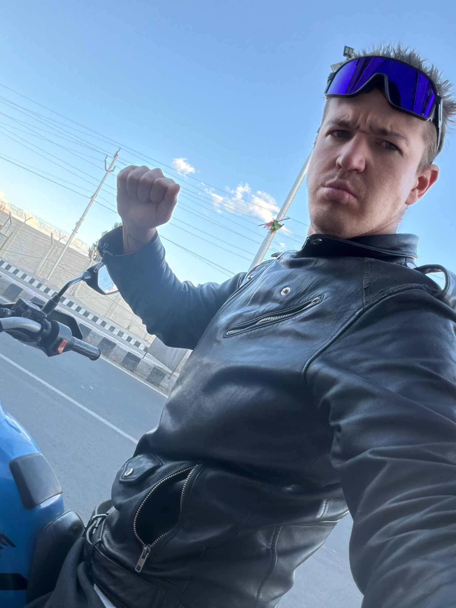 A man wearing a black leather jacket and blue goggles is seated on a blue motorcycle, captured in a selfie. He is raising his right fist in the air. The background shows a street in Leh, India, with overhead electrical wires and a barbed-wire fence. The sky is clear blue with some clouds. The man has a serious expression on his face. The image appears to have been taken during the daytime.