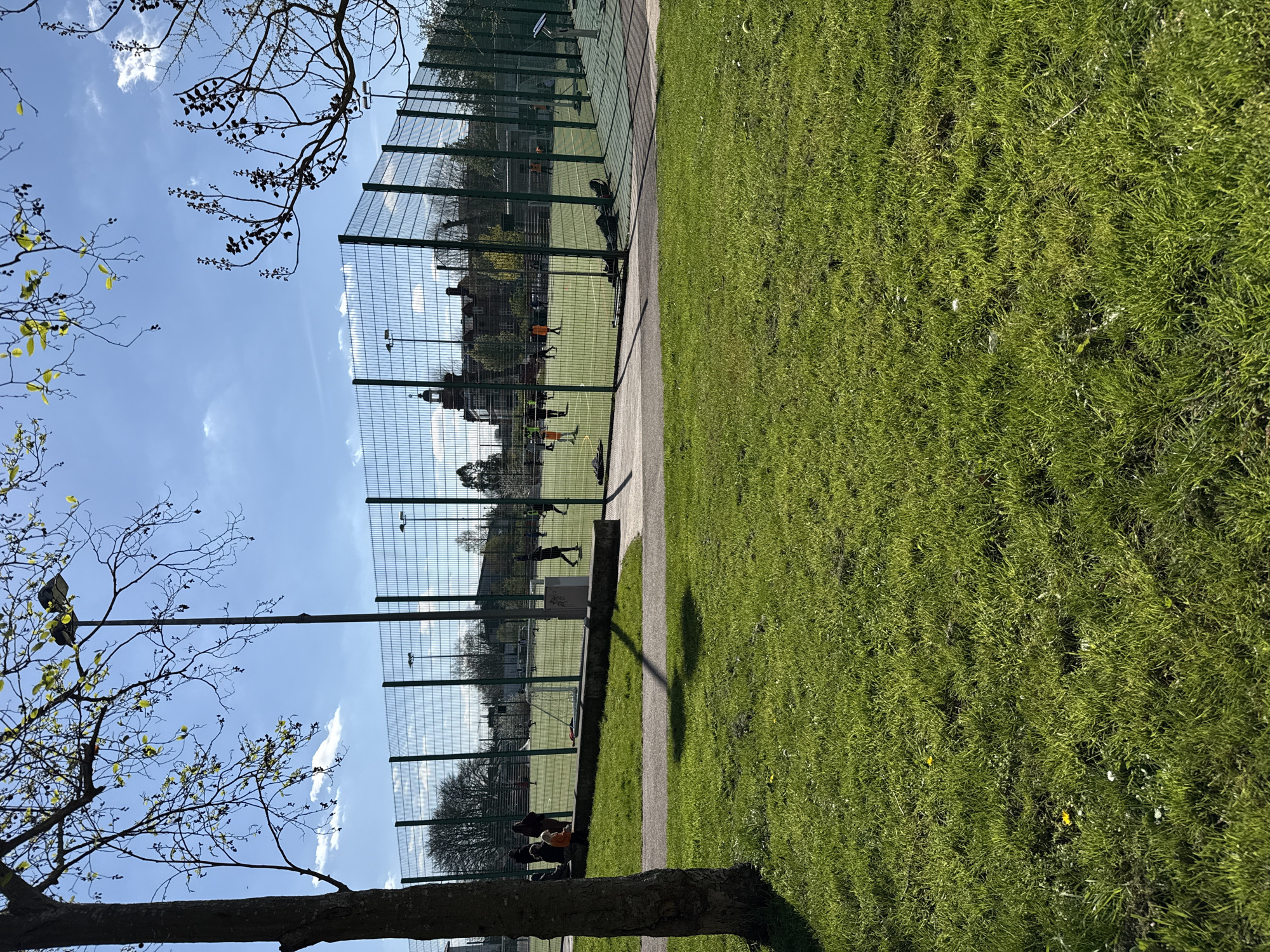 The image shows a public park in London, United Kingdom. A chain-link fence separates a green artificial turf sports field from a grassy lawn area. People are playing on the sports field. There is a paved walkway between the field and the lawn. Trees are visible, with branches partially obstructing the view in the foreground and background. The sky is blue with scattered clouds.
