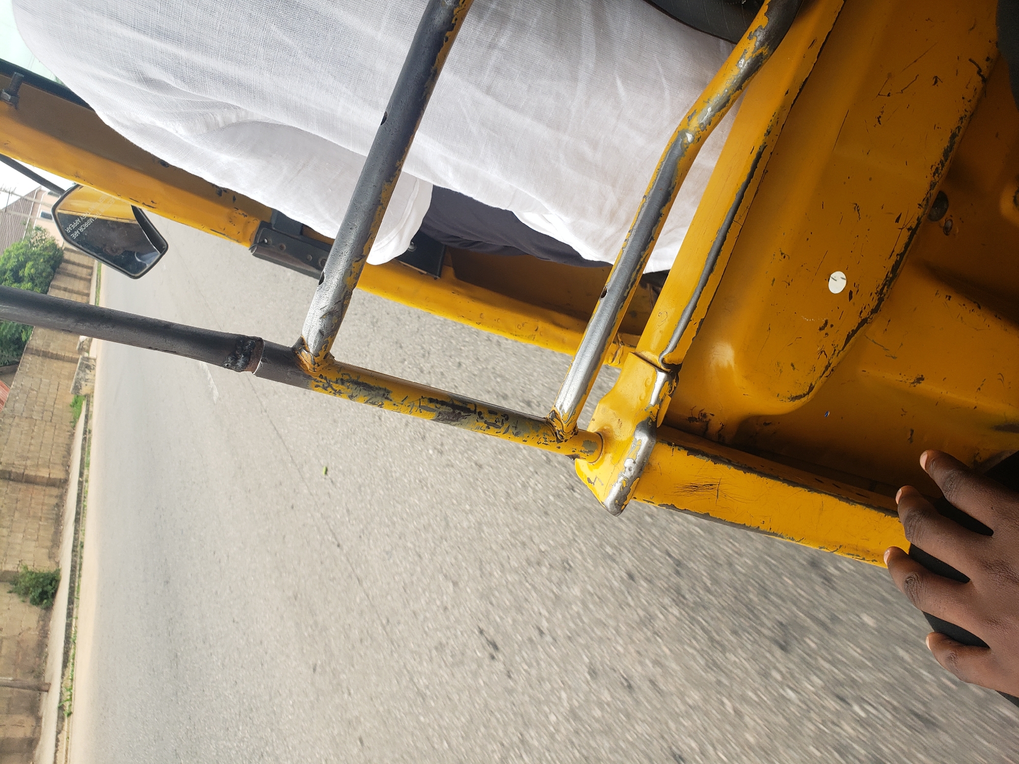 The image depicts a close-up view from inside a yellow motorized rickshaw, commonly known as an 'okada' or 'keke napep' in Nigeria. The perspective is from the passenger area looking towards the front and side. A person's dark-skinned hand is visible in the lower left corner, resting on a dark surface, possibly a seat or the body of the vehicle. The rickshaw's structure, made of yellow painted metal, is prominent, showing signs of wear and tear with peeling paint and rust in some areas. The interior includes a metal railing and a side mirror reflecting the surroundings.
The rickshaw is on a paved road, with motion blur indicating movement. The road surface is grey asphalt, showing some debris and texture. To the left of the road, a low wall made of concrete blocks lines the street. Beyond the wall, a sparse green bush is visible, and further in the background, buildings with terracotta roofs and trees can be seen, suggesting a suburban or urban environment. The sky is not visible in this framing. The context of Kaduna, Nigeria, suggests this scene likely takes place on one of the city's many roads, possibly in a residential or commercial area. The overall impression is of everyday transportation within the city.
