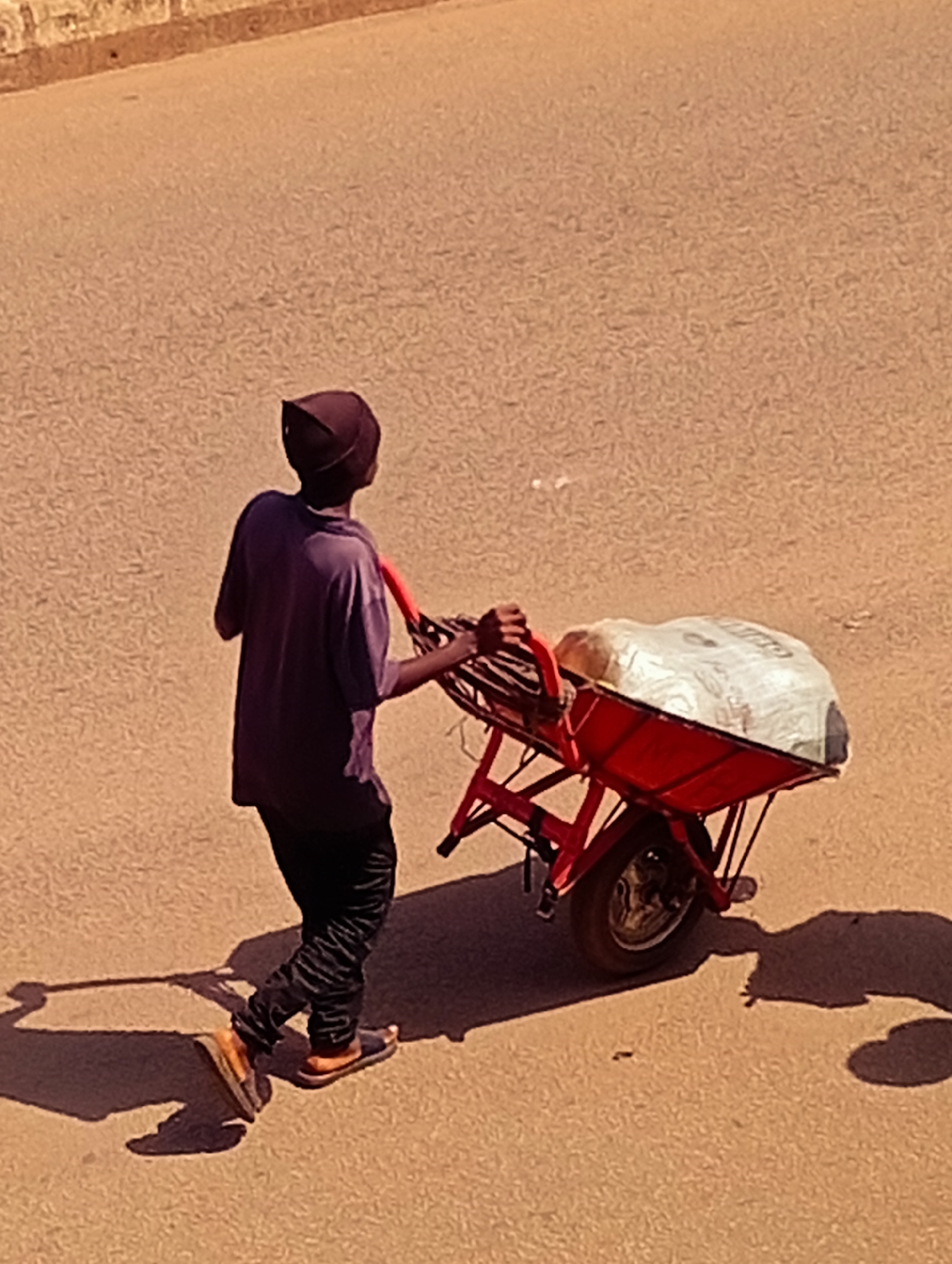 An individual, wearing a dark purple t-shirt, dark pants with a striped pattern, and sandals, is pushing a red wheelbarrow loaded with white material, possibly sacks or a tarp-covered object. The person is wearing a dark head covering. They are walking on a dusty, asphalt road in Bukuru, Nigeria. The road surface shows a slight incline. A shadow of the person and the wheelbarrow is cast on the road. The background consists of a textured, light-colored wall.