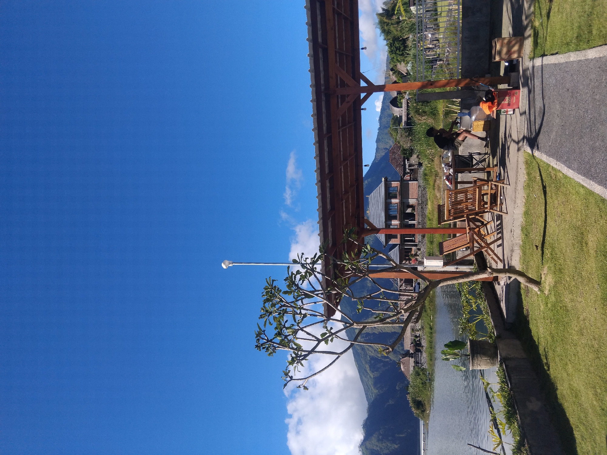 A landscape photograph, taken at an unknown time, shows a view from an unknown location in an unknown city. The scene is oriented vertically. A clear blue sky occupies much of the left side of the frame, punctuated by a few scattered white clouds.
A wooden structure with a roof runs diagonally across the upper portion of the image, casting a shadow over an area where people are present. To the right of the wooden structure, a man is seated on a chair, and appears to be interacting with items on a table or platform. A box with the word "Bintang" is visible nearby.
Wooden chairs and a table are arranged in front of the man. The ground is partly paved and partly covered with green grass. A body of water is below the wooden structure.
In the background, a mountain range is partially obscured by clouds. Various buildings and trees can be seen along the mountain slope. A tree with bare branches is near the lower left of the frame.