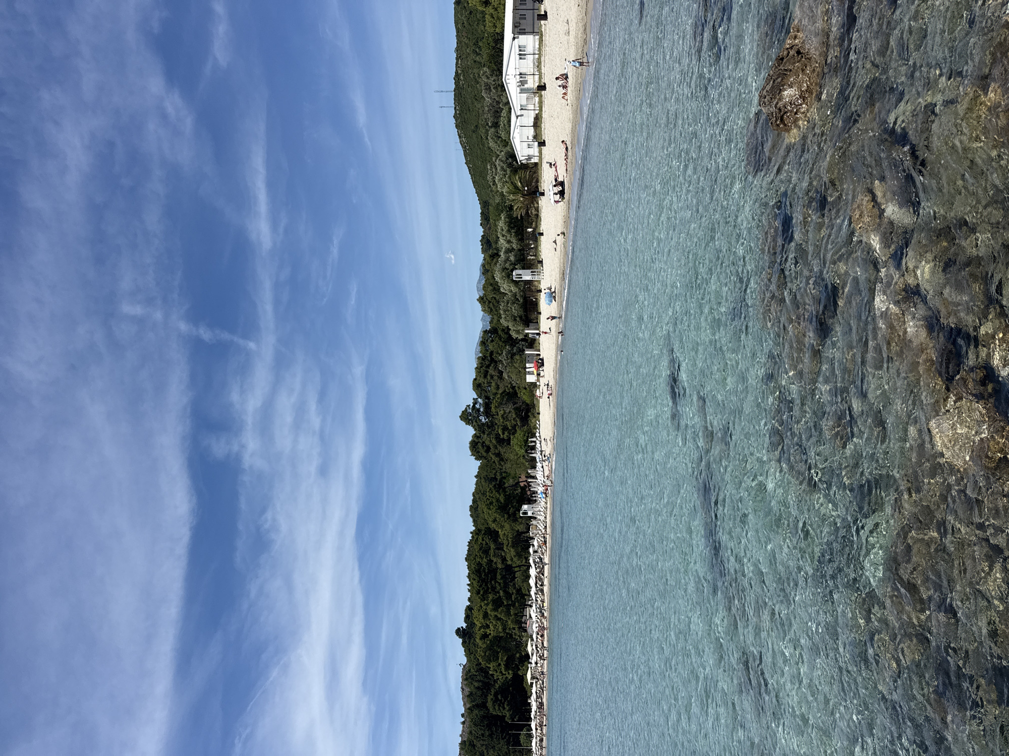 The image shows a coastline in Milovići, Montenegro, with clear turquoise water meeting a sandy beach. The water is relatively shallow near the shore, revealing submerged rocks and seabed. Along the beach, there is a line of trees and buildings, suggesting a resort or residential area. There are some people visible on the beach. The sky is blue with white clouds. The image is oriented with the sky to the left and the water to the right.
