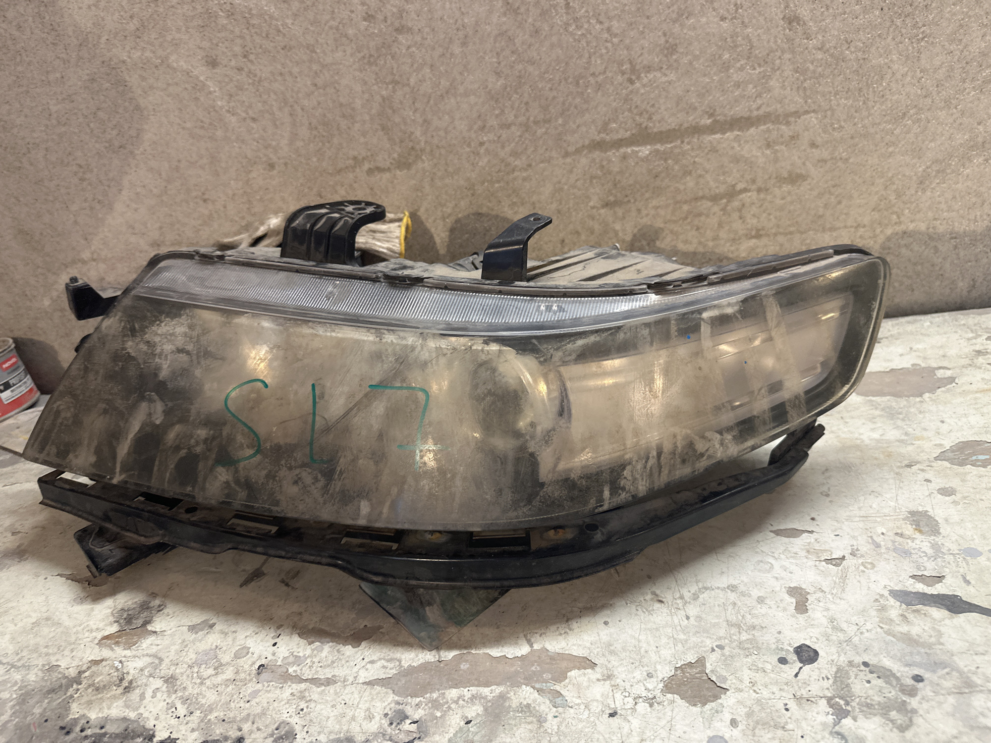 A dusty, used car headlight assembly sits on a speckled concrete surface in what appears to be a workshop or garage. The headlight is made of clear plastic and black plastic components, with visible scuffs, dirt, and what appears to be green marker writing on the lens that reads "SL 17". The background is a textured, light brown concrete wall. No people are visible in the image, and there is no information to indicate a specific location within International, Kyrgyzstan.
