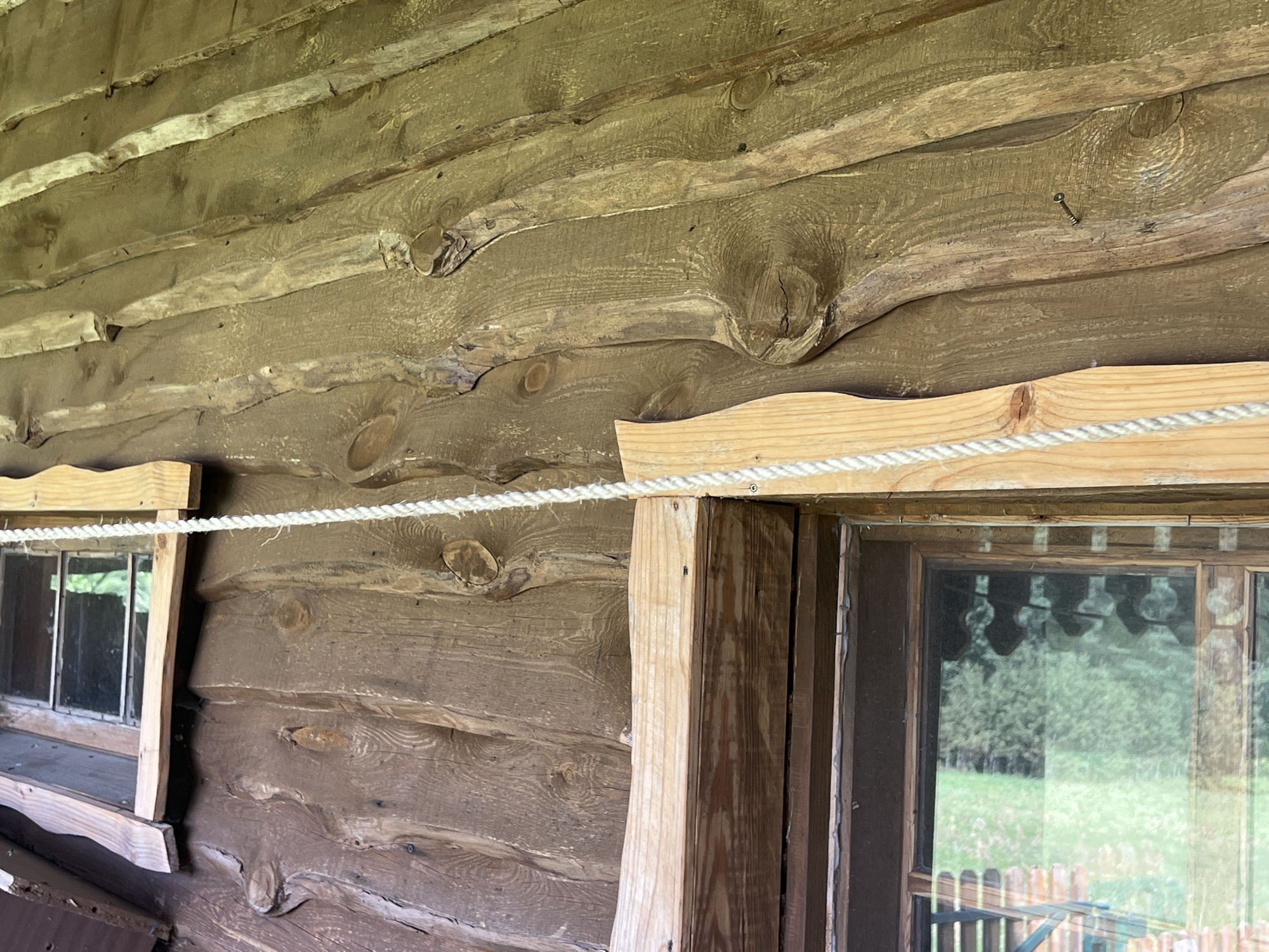 A close-up, eye-level shot shows the exterior wall of a rustic wooden building, possibly a historic structure in Norviliškės, Lithuania. The wall is constructed from rough-hewn logs, weathered to a dark brown hue, with visible knots and imperfections. Running horizontally across the frame, slightly above the center, is a thick, white rope, tautly stretched and secured to wooden elements above and around two windows.
The left window, partially visible, has a simple wooden frame and appears to be a small, possibly fixed pane. The right window is larger and shows a reflection of green trees and a wooden fence outside. The window frames and sills are constructed from lighter-colored wood, creating a contrast with the dark logs of the building. One of the wooden supports for the rope is a light-colored, unfinished plank, revealing its grain. Small screws are visible securing the rope and wooden trim. The overall impression is one of rural simplicity and aged construction.