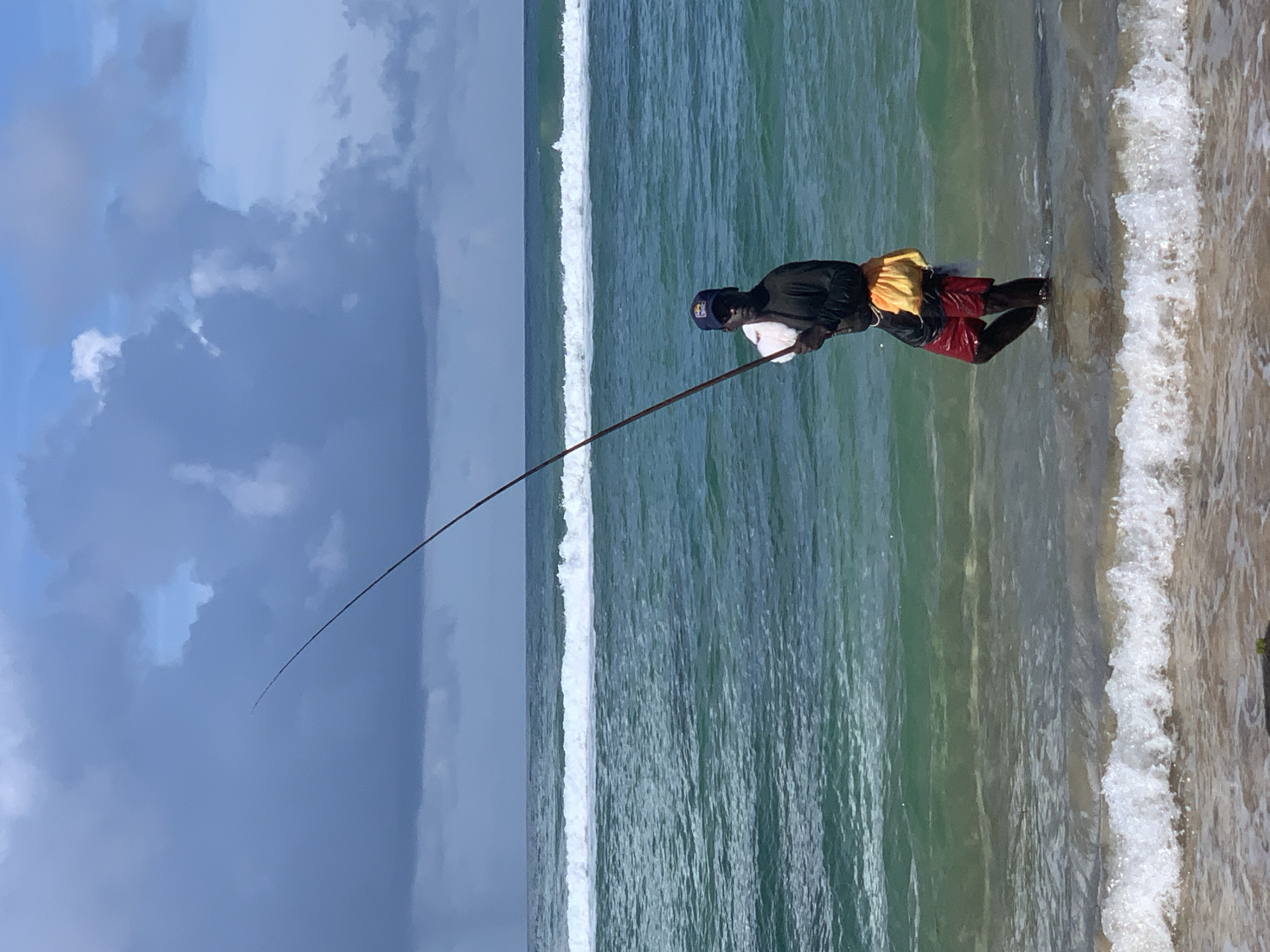 A man stands in the shallow ocean water, holding a long fishing rod that extends upward into the cloudy sky. He is wearing a dark long-sleeved shirt, red shorts, and a blue baseball cap. A white cloth is draped over his left shoulder, and a yellow pouch hangs from his right hip. Gentle waves break around his legs, and a wider wave line stretches across the horizon. The ocean water is a vibrant turquoise color, transitioning to a deeper blue further out. The sky is partly cloudy with patches of blue visible between dark gray clouds. The scene is set on the coast of Kathaluwa, Sri Lanka.