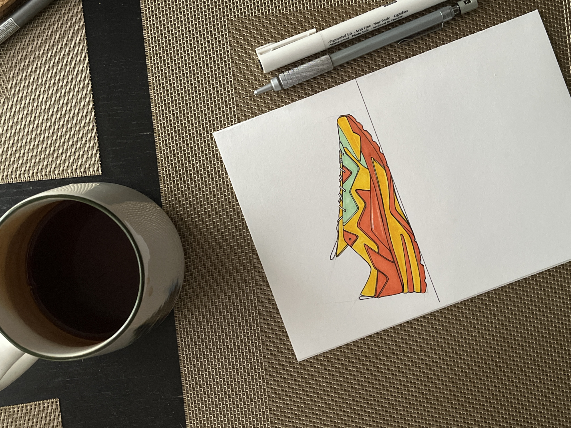 Here is a detailed description of the image:
**Scene:**
The image captures a still-life scene on a textured woven placemat, likely on a table.
**Objects:**
* **Cup:** A ceramic mug filled with a dark-colored liquid, possibly coffee or tea, is positioned in the lower left of the frame.
* **Drawing:** A white sketchbook page with a colorful drawing of an abstract object or design is in the upper right. The design features shades of red, orange, green, and yellow outlined in black.
* **Writing Utensils:** Two fine-liner pens (one white with a cap, one gray with a metallic tip) rest above the drawing, suggesting the drawing is in progress.
**Placement and Composition:**
The objects are arranged artfully, with the cup partially off-frame, drawing the viewer's eye across the scene from left to right. The drawing appears to be a work in progress, indicated by the presence of the pens.
**Environment:**
The background consists of the brown and tan woven placemat. The presence of the cup and drawing suggests this is a personal and informal setting.