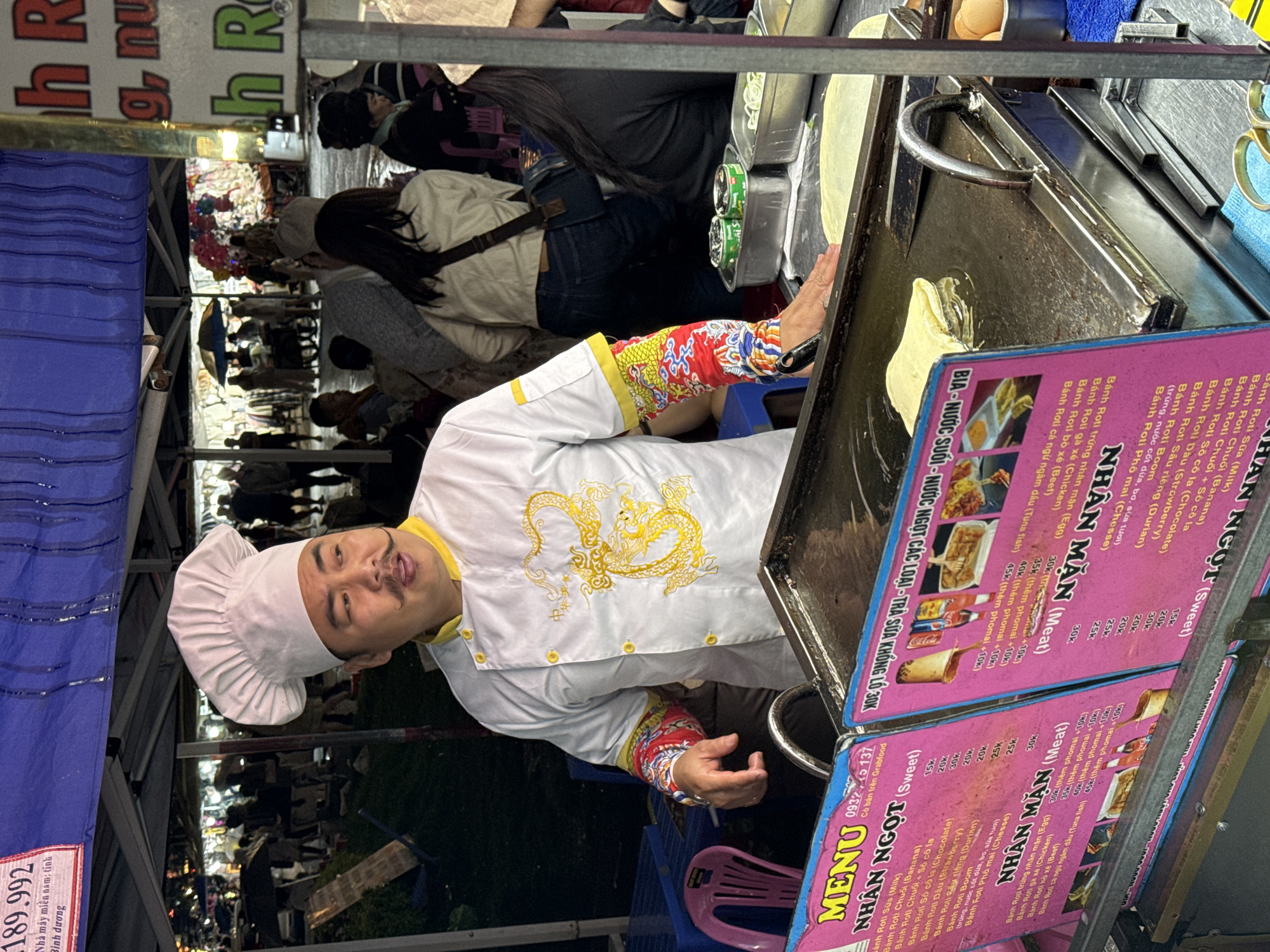 A photograph captures a man wearing a chef's hat and a white uniform with a golden dragon emblem. He is cooking at a food stall. The stall has a pink menu with Vietnamese text and food items listed. The man is near a hot cooking surface. The stall is set up in what appears to be a crowded outdoor setting. The general area has many people walking around, and there are tents or awnings overhead. A faded blue tarpaulin is visible in the top left, and behind the chef, people are milling about in what looks like a market or festival setting. Based on the menu featuring "Banh Roti", and the style of cooking, this is likely a street food stall in Da Lat, Vietnam.