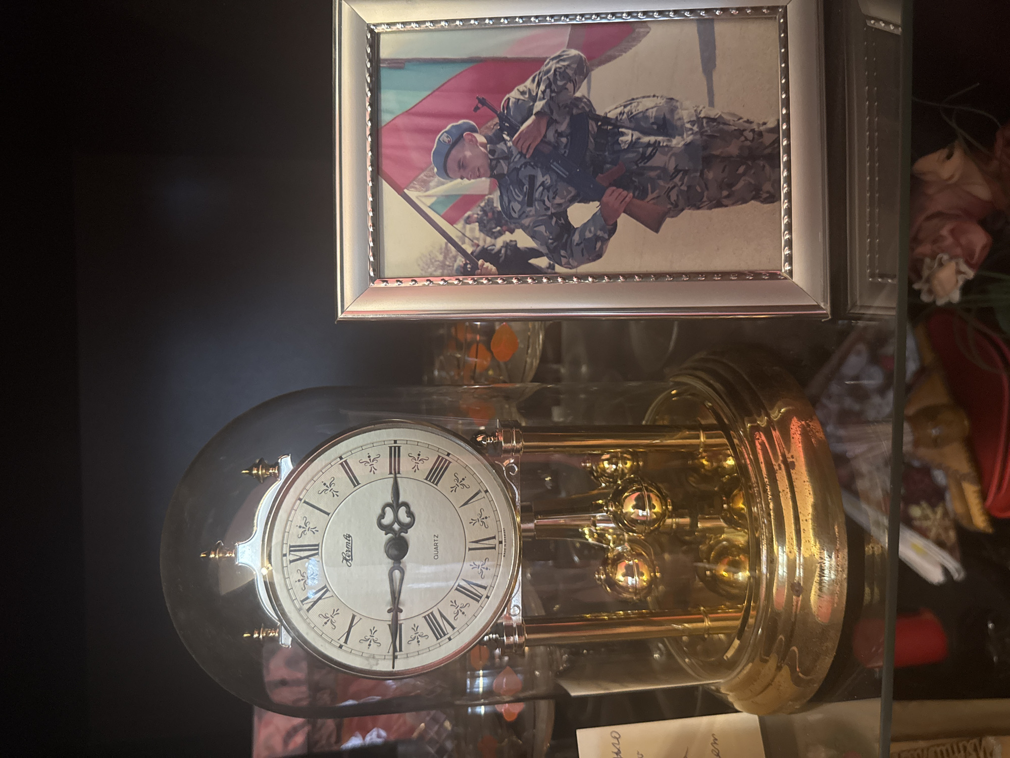 A photograph shows a framed picture and a clock under a glass dome. The picture, framed in silver, depicts a person in military uniform holding a rifle and standing in front of a flag with horizontal stripes of white, green, and red, possibly the flag of Bulgaria. The person is wearing a blue beret. The clock is a gold-colored anniversary clock with a white face and black Roman numerals. The clock's hands indicate the time is approximately 6:06. The objects are situated on a dark surface, possibly a table or shelf, with other items scattered around, including what appears to be a candy dish. The city of Burgas, Bulgaria is not visible in the image and cannot be determined from the available information.