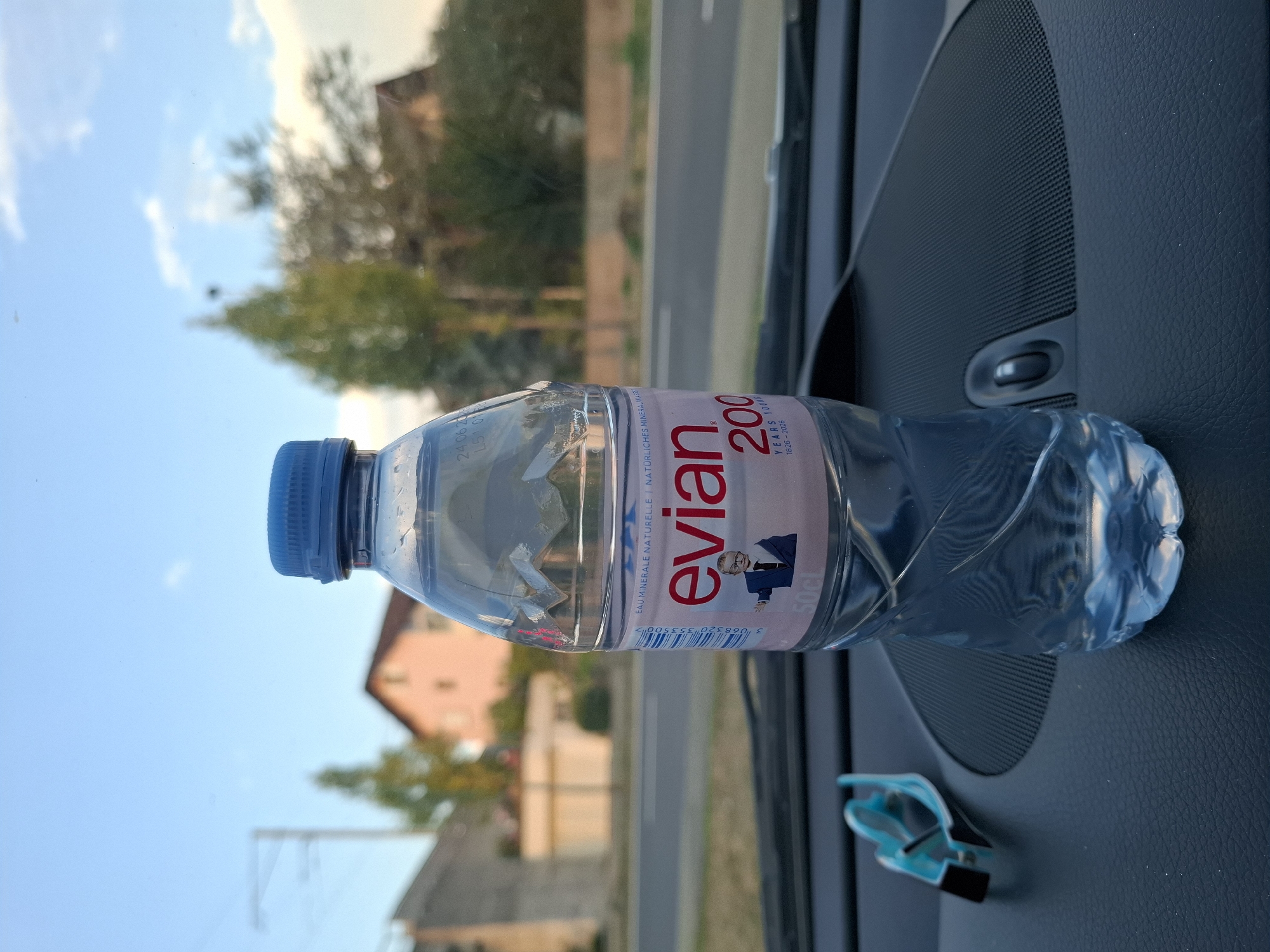 A close-up, eye-level shot shows a 50cl bottle of Evian water with a blue cap, standing on the dashboard of a car. The bottle is partially filled with water and has a label indicating "Evian 200 YEARS YOUNG 1826-2026". To the left of the bottle, a blue and black clip is visible. The dashboard is made of black textured material. In the background, out of focus, is a street with a road and sidewalk, flanked by houses and trees. The sky is visible above, with some clouds. The scene appears to be located in Hochdorf, Switzerland.