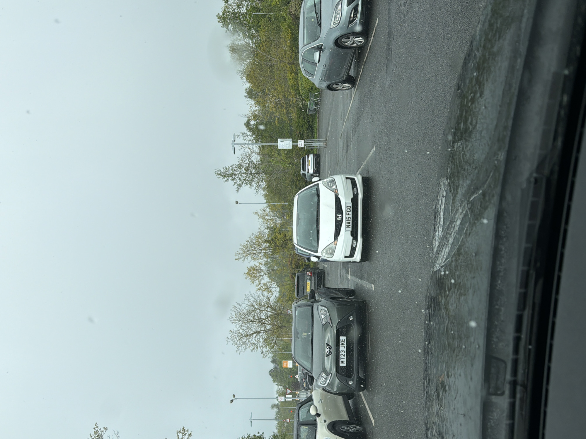 The image shows a parking lot in Pity Me, United Kingdom on a rainy day. Multiple cars are parked in rows. A white Honda with the license plate "NA15 FGO" is prominently visible. Behind it is a dark car and in front of the Honda is a grey Toyota with the license plate "MT23 JKE". Other vehicles are also parked nearby. The trees in the background suggest it is near a wooded area. The image was taken from inside a car.