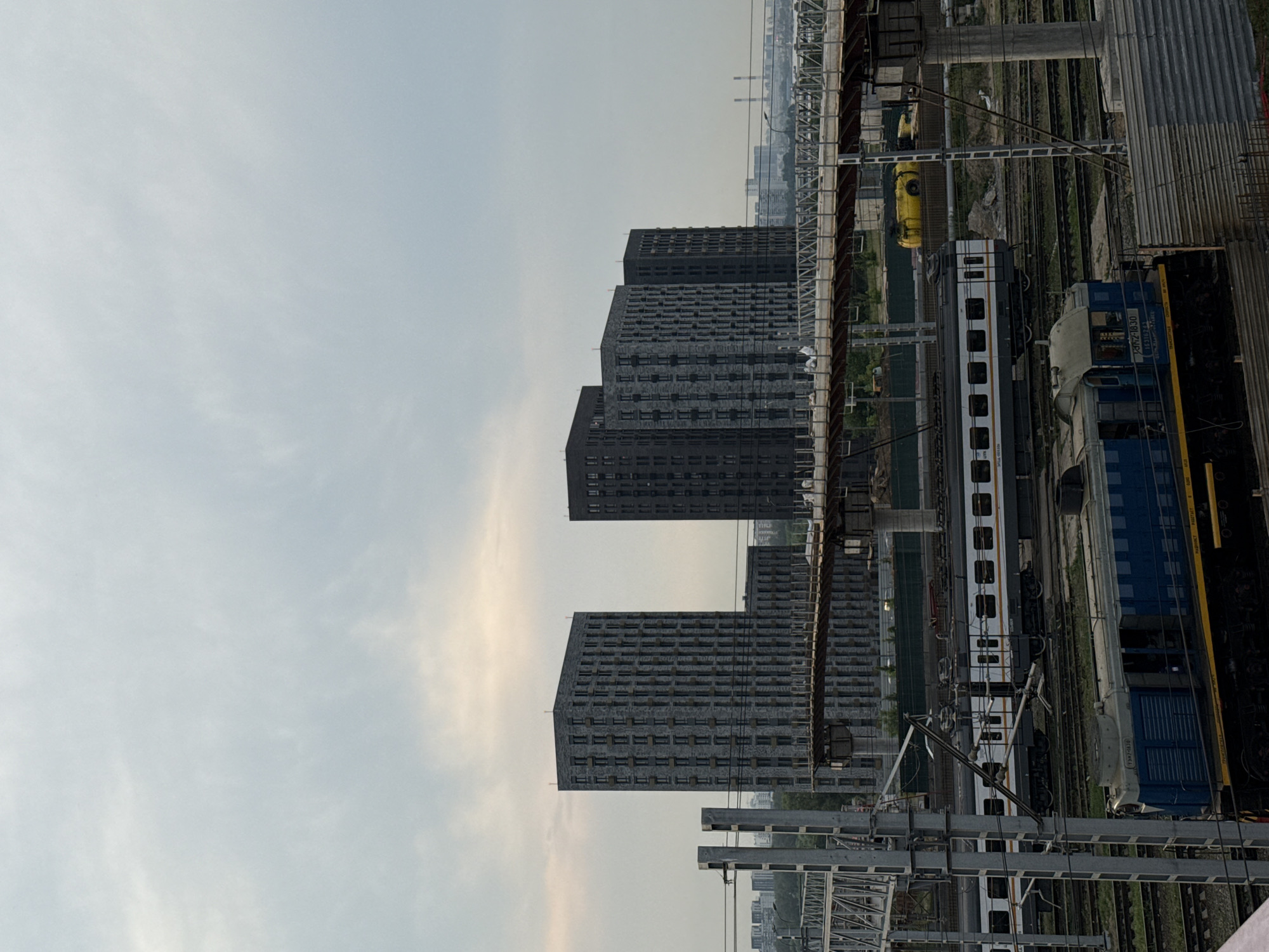 The image shows a view of a construction site with several buildings and trains in Moscow, Russia. In the background, four tall, dark-colored residential buildings are visible. In the foreground, a blue and yellow train engine is parked on the tracks, with another white and gray passenger train behind it. A bridge structure is under construction above the tracks, with yellow construction equipment near it. The sky is overcast, with some hints of sunlight breaking through. The overall scene suggests an urban environment with ongoing development.