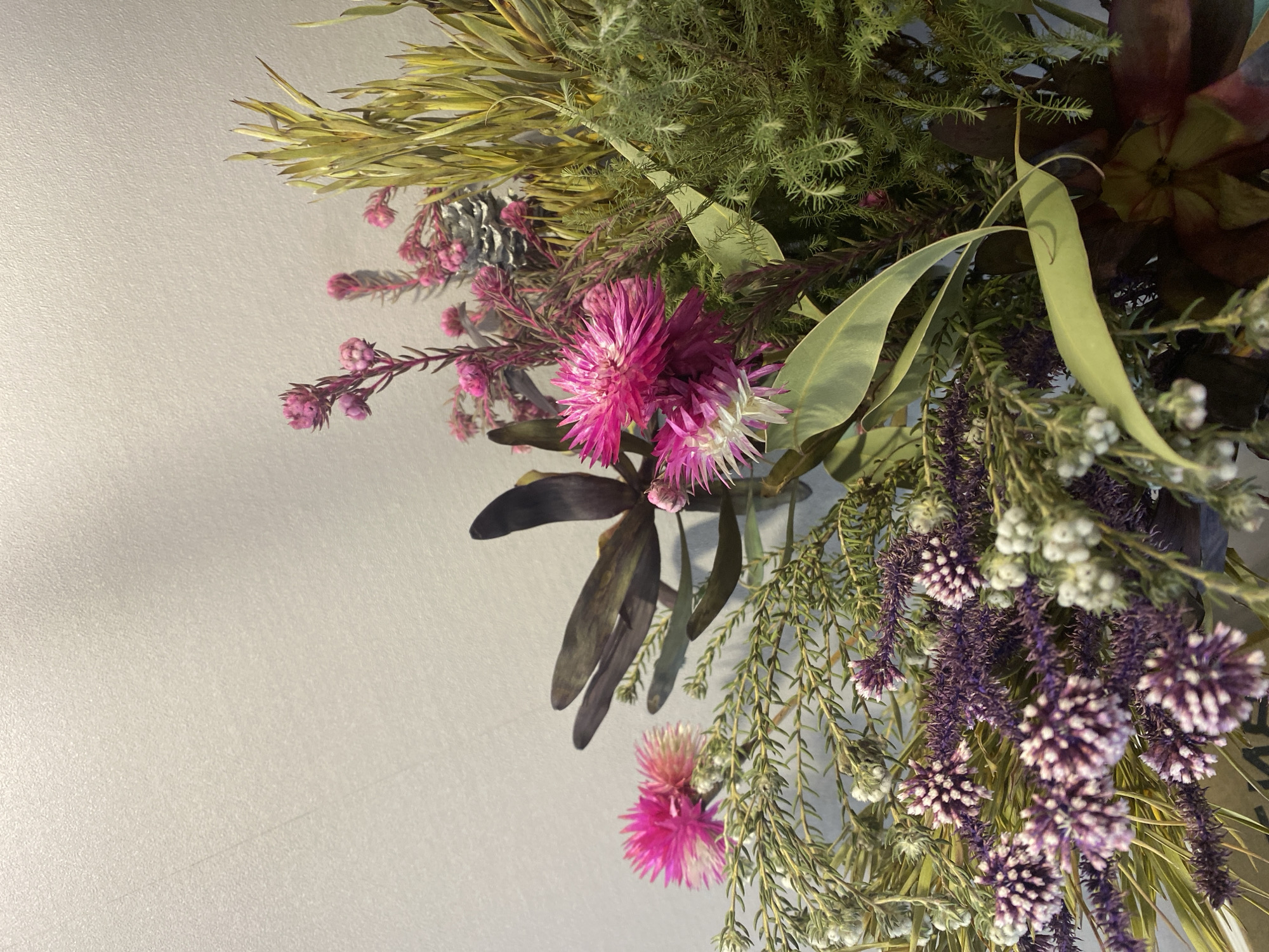 A close-up shot of a dried flower arrangement. The arrangement contains a variety of dried plants, including yellow foliage, purple spherical flowers, pink tassel-like flowers, and eucalyptus leaves. The background is a light grey or white wall. The location is not evident from the image. There is no information available to assume the image was taken in the city of Chaikovsky, Russia.