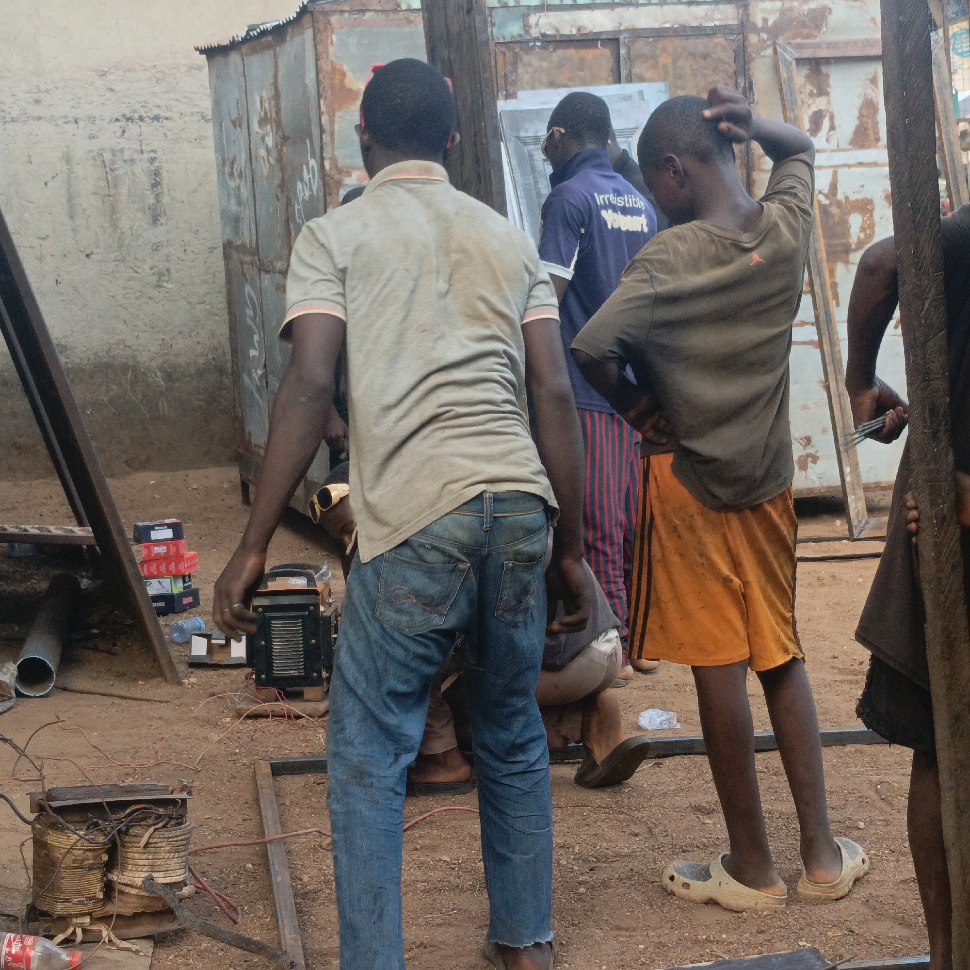 The image depicts a scene in Akwanga, Nigeria, seemingly at a metal workshop. Several men and a boy are visible. One man stands with his back to the camera, wearing a light-colored t-shirt and blue jeans. Another man in a blue "Irresistible Yobourt" t-shirt stands nearby. A boy in a brown shirt and orange shorts is also present, scratching his head. An additional individual is partly visible on the right, holding a tool.
An electric generator sits on the ground alongside a pile of boxes. A used transformer is also on the ground with exposed wires. Metal sheets and frames are scattered around, suggesting the environment of a workshop. The ground is dirt. A partially obscured structure made of corrugated metal is visible in the background.