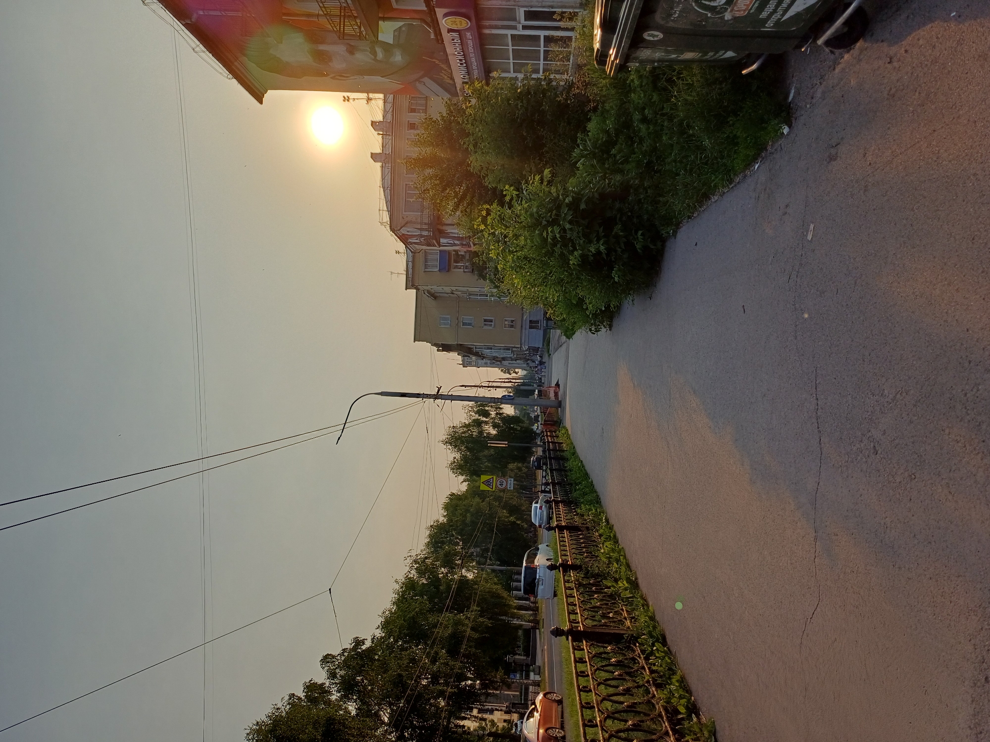 A street scene in an unknown city during sunset. The sun is low in the sky, casting a warm glow. A sidewalk runs along the left side of the image, bordered by a decorative metal fence and lush green grass. Beyond the fence, cars are driving on a road lined with trees. On the right side of the sidewalk, there is a large building with a mural of a man's face on its side. Further down the street, there are more buildings, some with balconies, and more trees. Two large green trash bins are visible on the right edge of the image. The sky is clear with a few faint clouds. Overhead, power lines are strung across the sky.