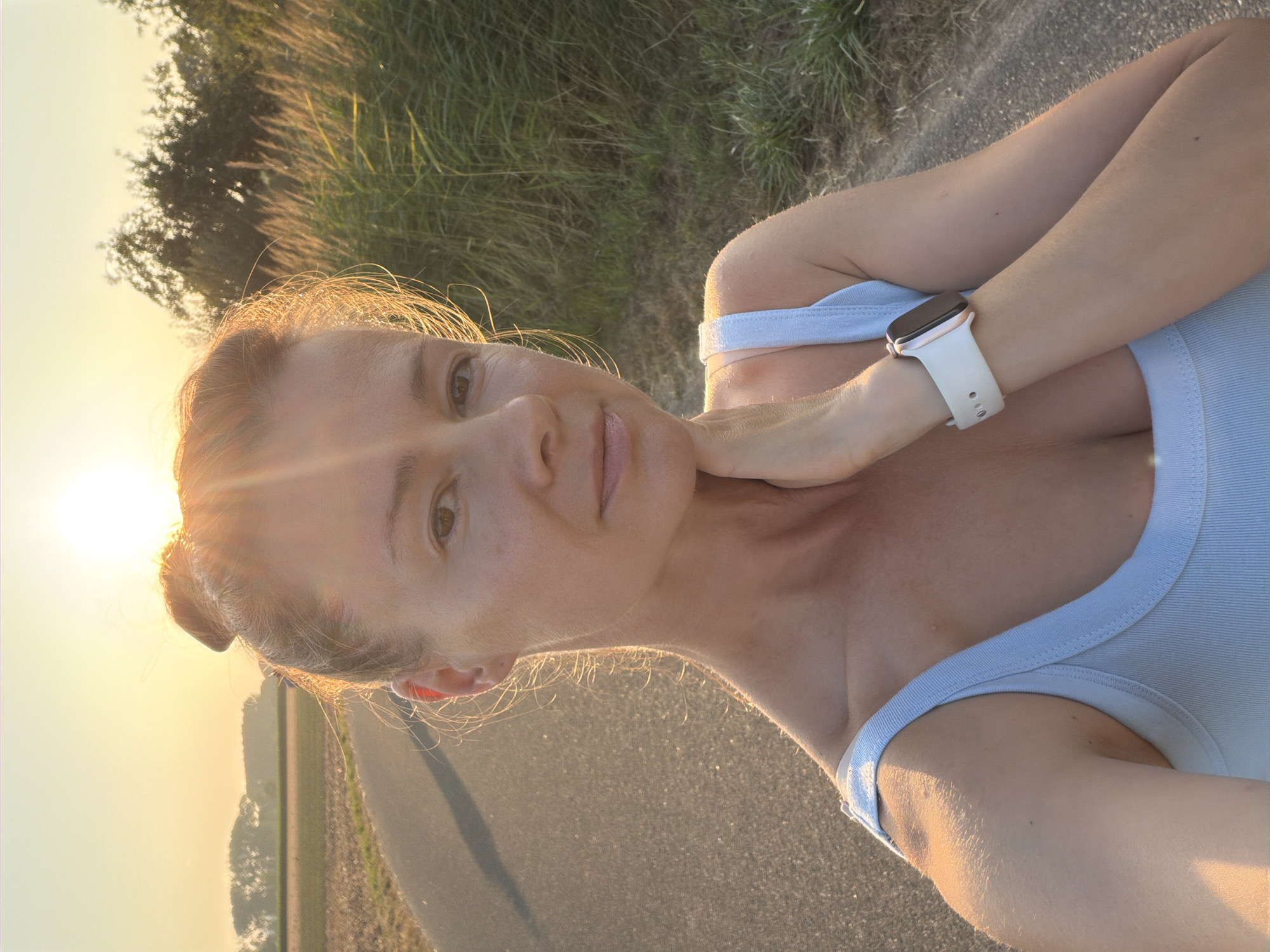 A middle-aged Caucasian woman is captured in a selfie outdoors, standing on a paved path with fields and vegetation on either side. The sun is low in the sky, casting a warm golden light that illuminates her face and hair. She has her hair styled in a bun on top of her head, and her left hand is resting on her neck. She is wearing a light blue tank top and a white smartwatch on her left wrist. The background suggests a rural or semi-rural setting, possibly on the outskirts of Melick, Netherlands, given the presence of fields and tall grasses typical of the Dutch landscape. The overall scene conveys a sense of calm and possibly post-exercise activity, with the warm sunset creating a serene atmosphere.