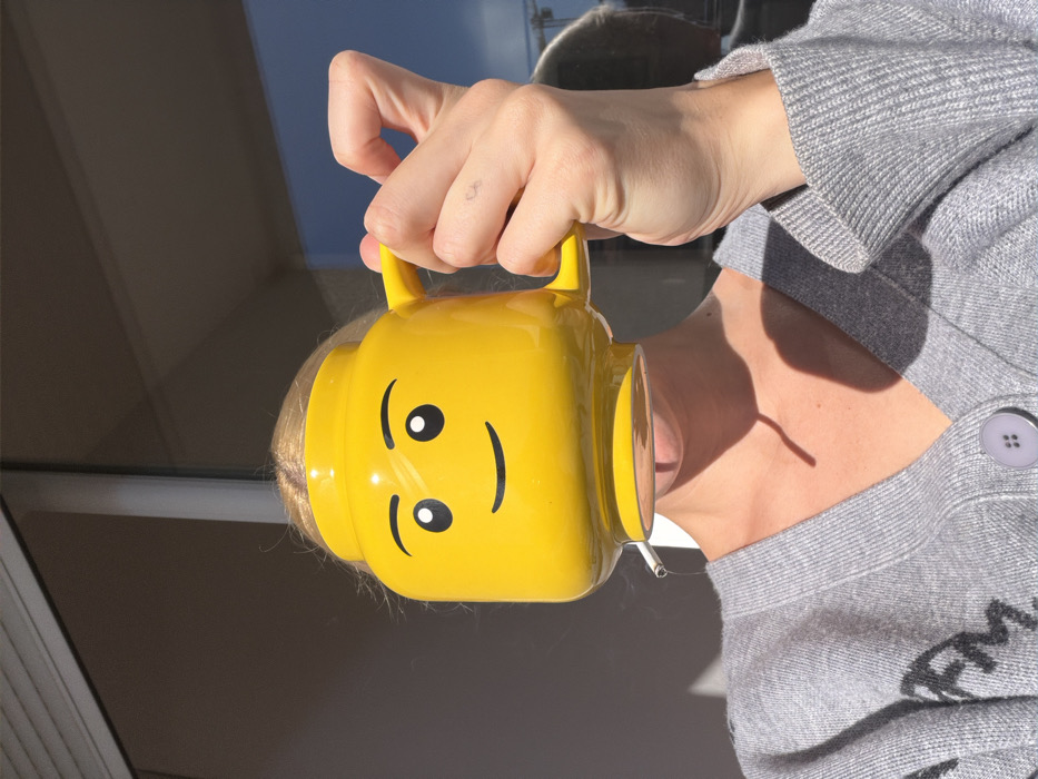 A partially visible individual is depicted outdoors in Budva, Montenegro, under direct sunlight. The subject, possessing blonde hair, wears a light grey V-neck knit cardigan with a visible light purple button and partially obscured dark lettering on the left chest area. A bright yellow, ceramic mug, designed to resemble a LEGO minifigure head with black eyes and a smiling mouth, is held by the subject's right hand, obscuring most of their face. A lit cigarette is held between the subject's lips, protruding from beneath the mug's base, with a wispy trail of smoke rising to the left. A small, dark tattoo is visible on the lateral side of the subject's right index finger. The background features a large, reflective glass surface, likely a window or balcony door, which mirrors a clear blue sky and a distant construction crane structure. Strong illumination casts distinct shadows from the mug onto the subject's neck and upper chest.