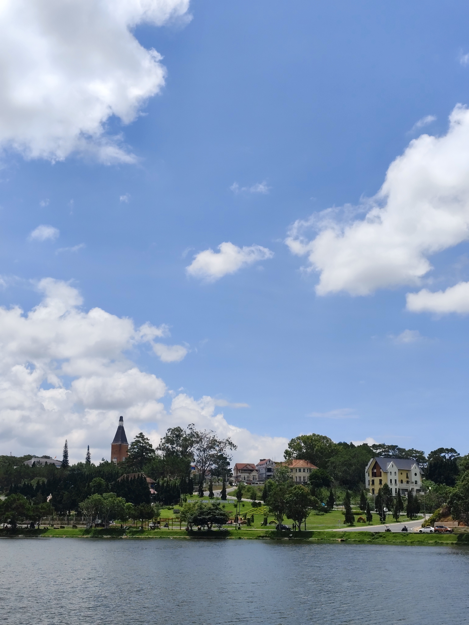 The image depicts a serene view of Xuan Huong Lake in Da Lat, Vietnam. The lake's dark blue waters stretch across the lower portion of the image. In the background, a landscape rises with lush green trees and a manicured grassy area, indicating a park or garden setting. Architectural elements, including a distinctive tower and several buildings with varying architectural styles and colors, are visible among the trees. A road runs along the edge of the park, adjacent to the lake, with a few vehicles parked or moving along it. The sky dominates the upper half of the image, featuring a vivid blue expanse with scattered, puffy white clouds.