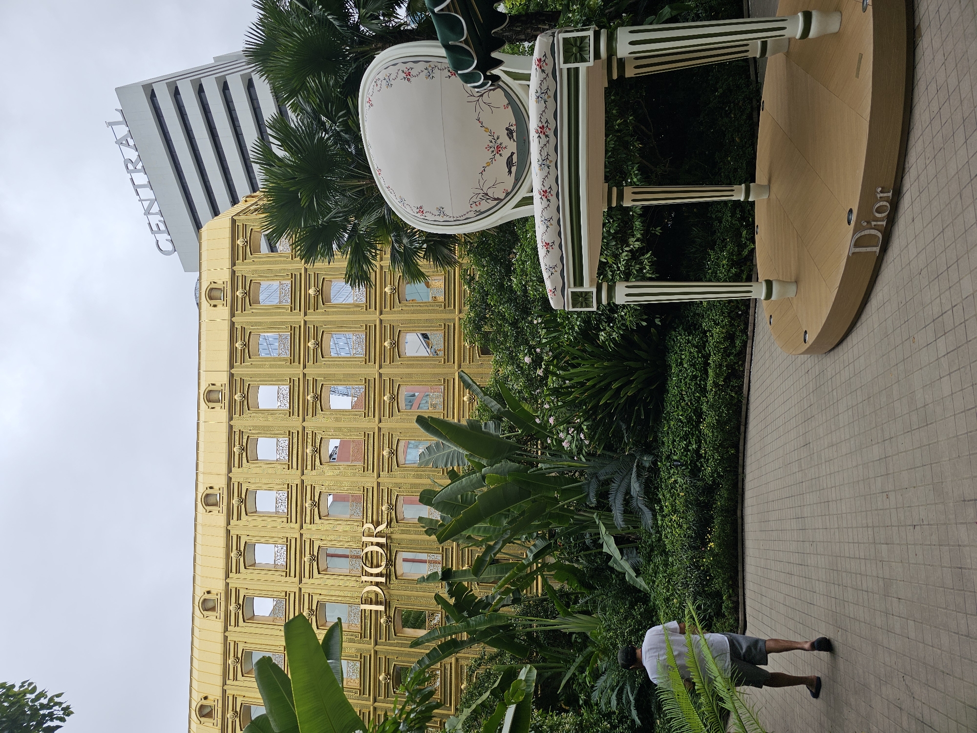 The image depicts the Dior store in Bangkok, Thailand, located in the Siam Paragon shopping mall. A man in a white t-shirt and grey shorts is walking away from the camera on a paved walkway. In the foreground, there is a large, decorative chair with floral patterns, positioned on a circular wooden platform that bears the "Dior" logo. Behind the chair and walkway, lush greenery and tropical plants are visible. The Dior store itself is a prominent golden building with numerous windows. In the background, a white building with the "CENTRAL" logo is visible. The sky is overcast.