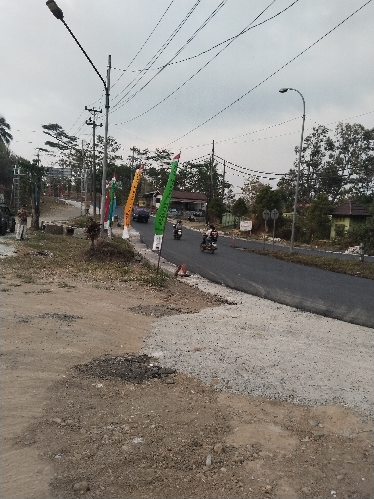 The image depicts a street scene in Kalibakung, Indonesia, with a gray, overcast sky. The foreground is a dirt and gravel area with patches of asphalt, suggesting ongoing road construction or repair. A paved road runs diagonally from the mid-ground to the right. Along the side of the road, there are several colorful pennants or flags, some with Indonesian writing, including one that reads "DESA WISATA KALIBANUS." A street lamp is visible on the left side of the road, and another on the right. In the mid-ground, several trees and buildings line the road. Two individuals are on motorcycles, one traveling on the paved road and another further down the road. A vehicle is parked on the left side of the road. The overall impression is of a semi-rural or suburban area undergoing development, with a mix of natural and man-made elements.