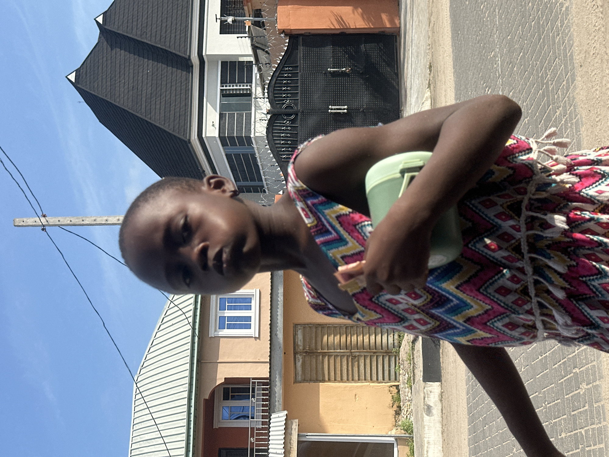 A young Black child, with dark skin and short hair, is pictured outdoors in Badagry, Nigeria, on a sunny day. The child is facing the camera and is holding a light green container and a piece of food. The child is wearing a colorful, sleeveless dress with a geometric pattern in shades of pink, yellow, blue, and white, and features white tassels around the waist.
In the background, there are residential buildings and a large black metal gate with spiked ornamentation. The building on the left has a peach-colored facade with brown shutters and a white corrugated roof. The building behind the child features a dark, multi-peaked roof. The ground is paved with interlocking gray bricks. A concrete pole with electrical wires stretches across the upper portion of the image.