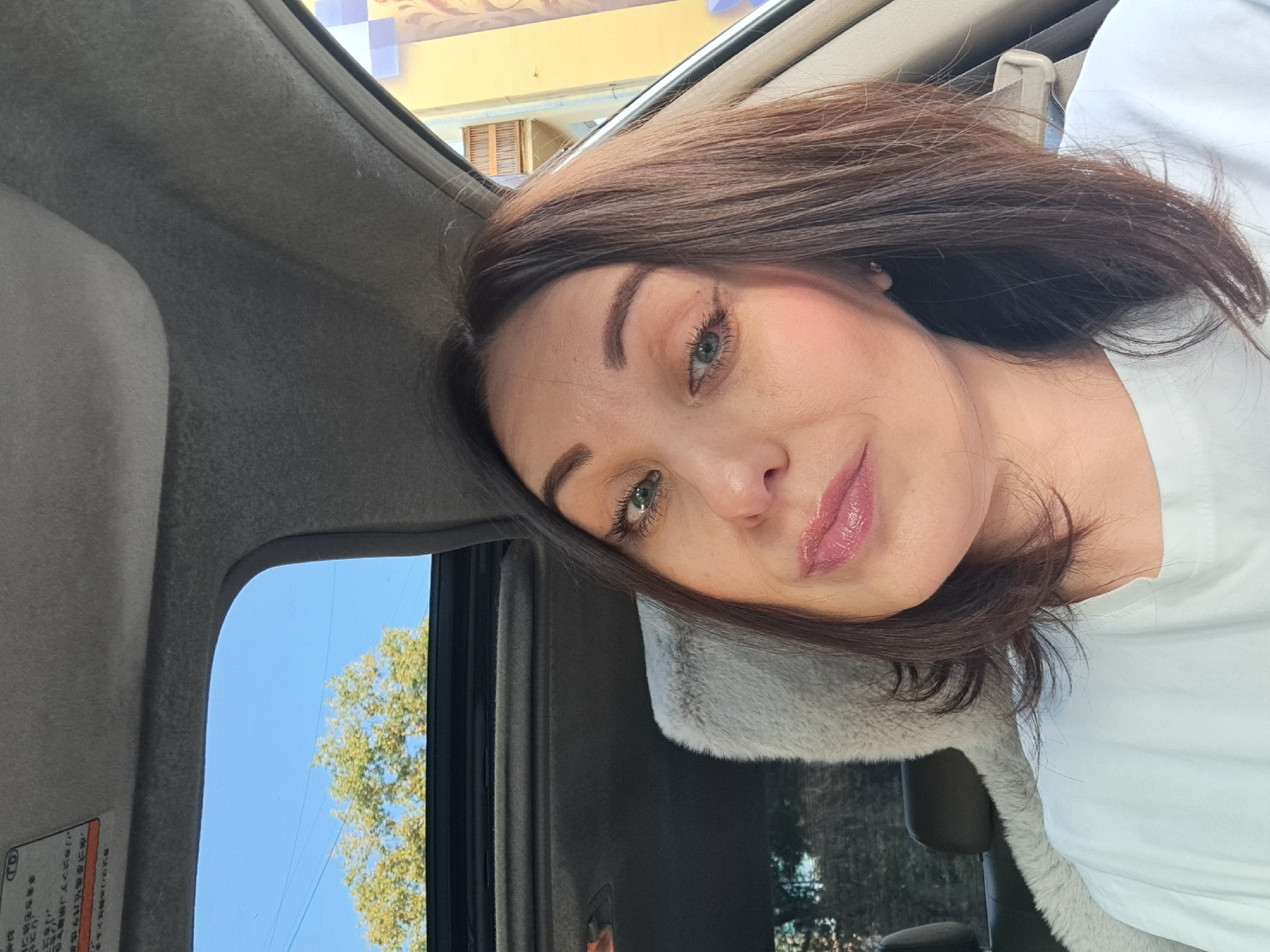 A woman with shoulder-length brown hair and green eyes is taking a selfie inside a car. She is wearing a white t-shirt. The car has a sunroof open, revealing a blue sky and the top of a leafy green tree. A fuzzy grey seat cover is visible. In the background, a yellow building with balconies can be seen. There are some faint markings on the car's interior ceiling. The scene is likely set in Irkutsk, Russia, given the context provided.