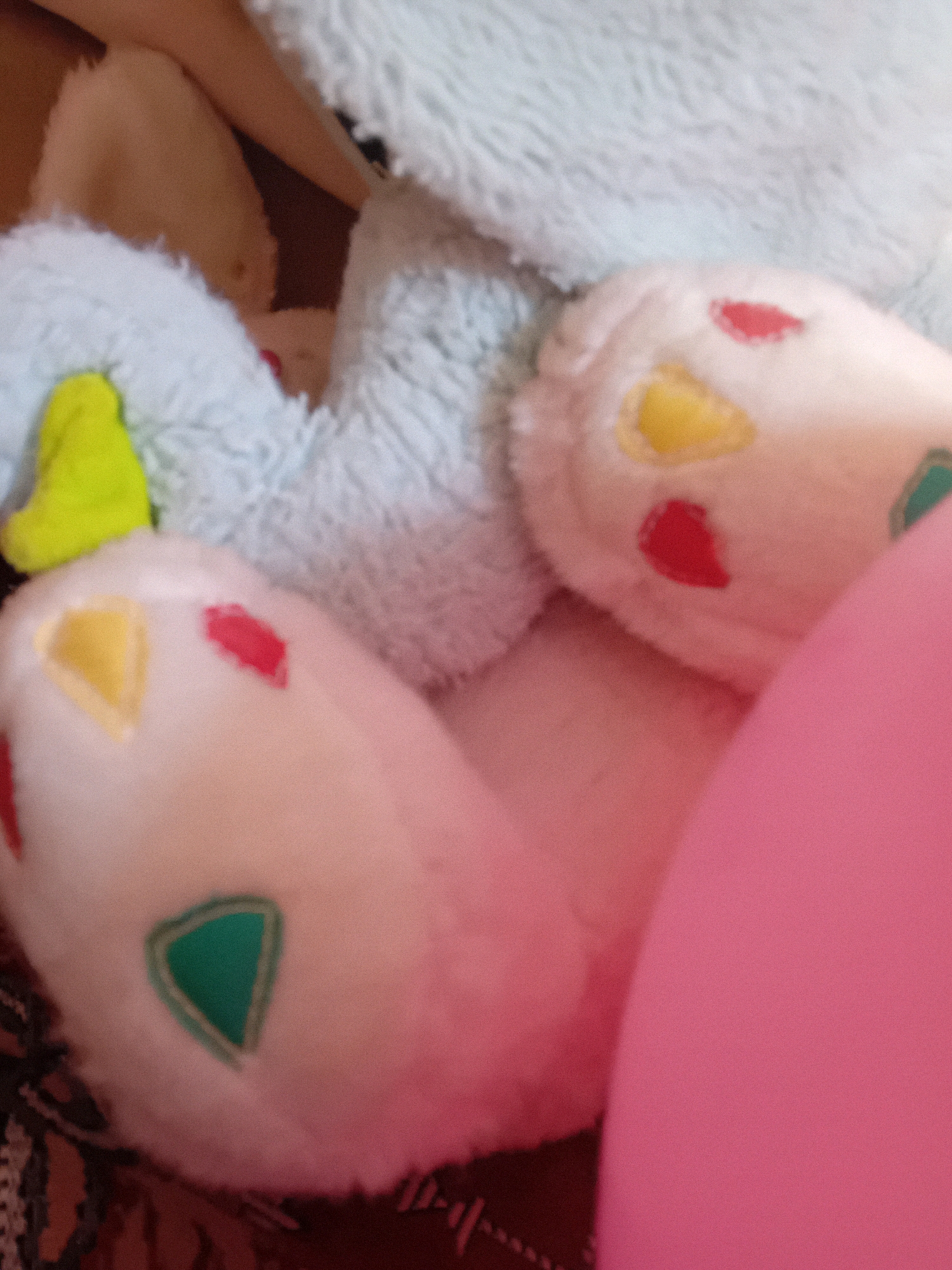 The image depicts a close-up view of plush toys. In the foreground, there are two white, foot-like appendages with colorful gem-shaped embellishments: a yellow teardrop shape and a red teardrop shape on one, and a yellow teardrop shape and a green triangle on the other. These appendages are part of a larger, light blue plush toy, visible in the background, which has a fuzzy texture. A large, bright pink object, possibly another plush toy or a cushion, dominates the right side of the frame. The overall lighting is soft, creating a slightly blurred effect. The scene is indoors, with a hint of a patterned surface visible at the bottom left, suggesting it is on a floor or rug. There is no identifiable location within the city of Chuhuiv, Ukraine, or any specific context provided by the image itself.