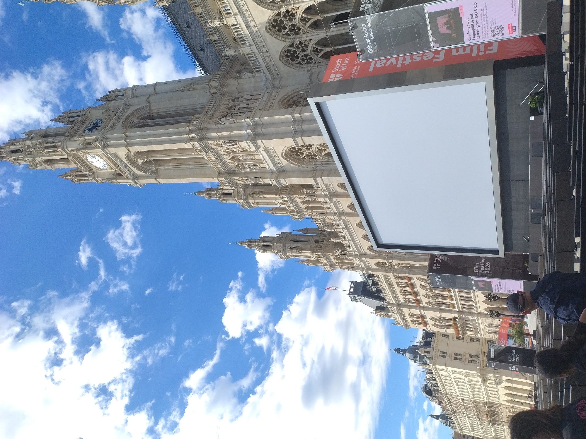The image depicts an outdoor cinematic event held in front of the Vienna City Hall (Rathaus Wien) in Vienna, Austria. A large, blank white screen is positioned centrally, suggesting an outdoor movie screening or event. In the background, the ornate Gothic architecture of the Rathaus Wien is prominently visible, with its distinctive clock tower and intricate facade. Several tall banners are placed on the right side of the screen, one of which features the text "Film Festival" vertically. To the left of the screen, the continuous facade of the Rathaus Wien extends, featuring numerous windows and decorative elements. In the foreground, rows of empty dark seats are arranged, indicating the seating area for attendees. A few individuals are partially visible in the lower portion of the frame, with their backs to the camera, suggesting they are part of the audience or staff. The sky above is a bright blue, dotted with fluffy white clouds, indicating fair weather. The overall scene conveys the atmosphere of a public event taking place in a historically significant urban setting.