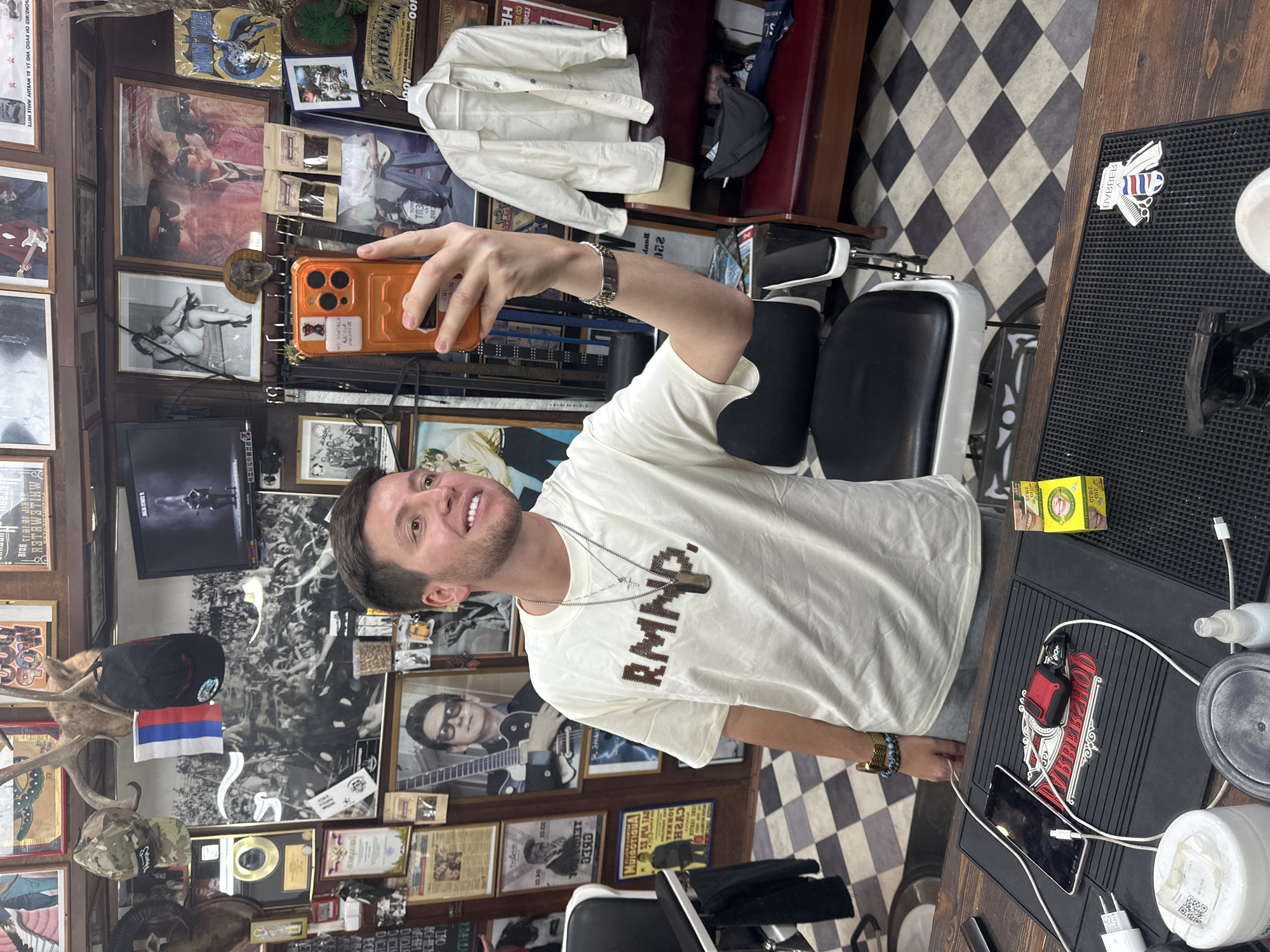 A man is taking a selfie in a barber shop. He is holding an orange phone case and wearing a white T-shirt with "RMND" printed on it. Behind him is a wall covered in photos, posters, and other decorations. There's a barber chair next to him and a black mat with barber tools. The floor has a black and white checkered pattern. The barber shop is cluttered with items and has a retro feel. The location of the barber shop is Moscow, Russia.
