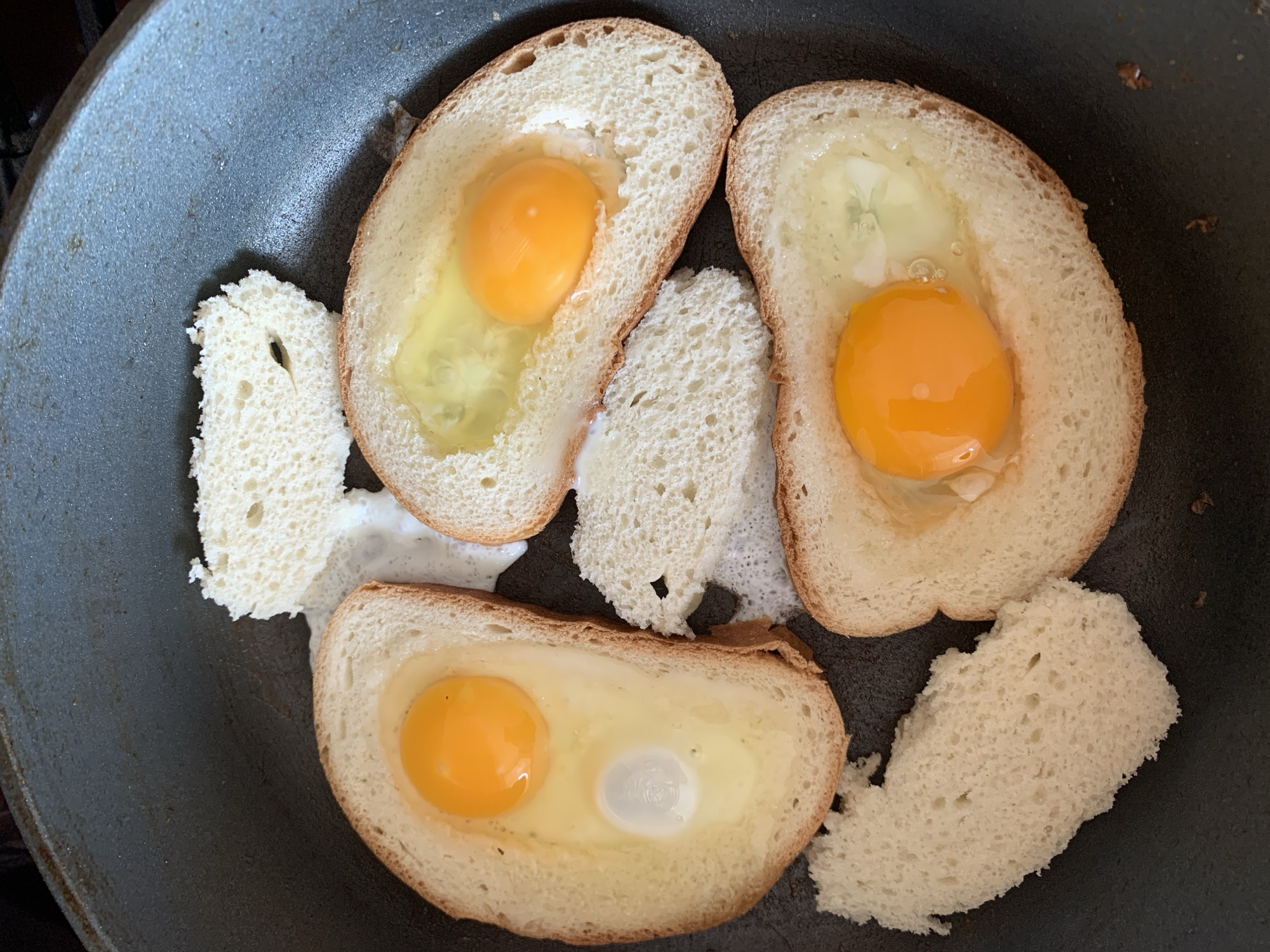 A close-up, overhead shot shows three slices of white bread, each with a hole in the center, cooking in a black non-stick frying pan. One egg yolk and some egg white are visible in each of the bread holes. Two small pieces of bread are also in the pan. The scene is an indoor kitchen, likely in Kapyĺ, Belarus, with natural light. The eggs appear to be partially cooked, with the yolks still distinct and the whites beginning to set. The bread is lightly browned on the edges.