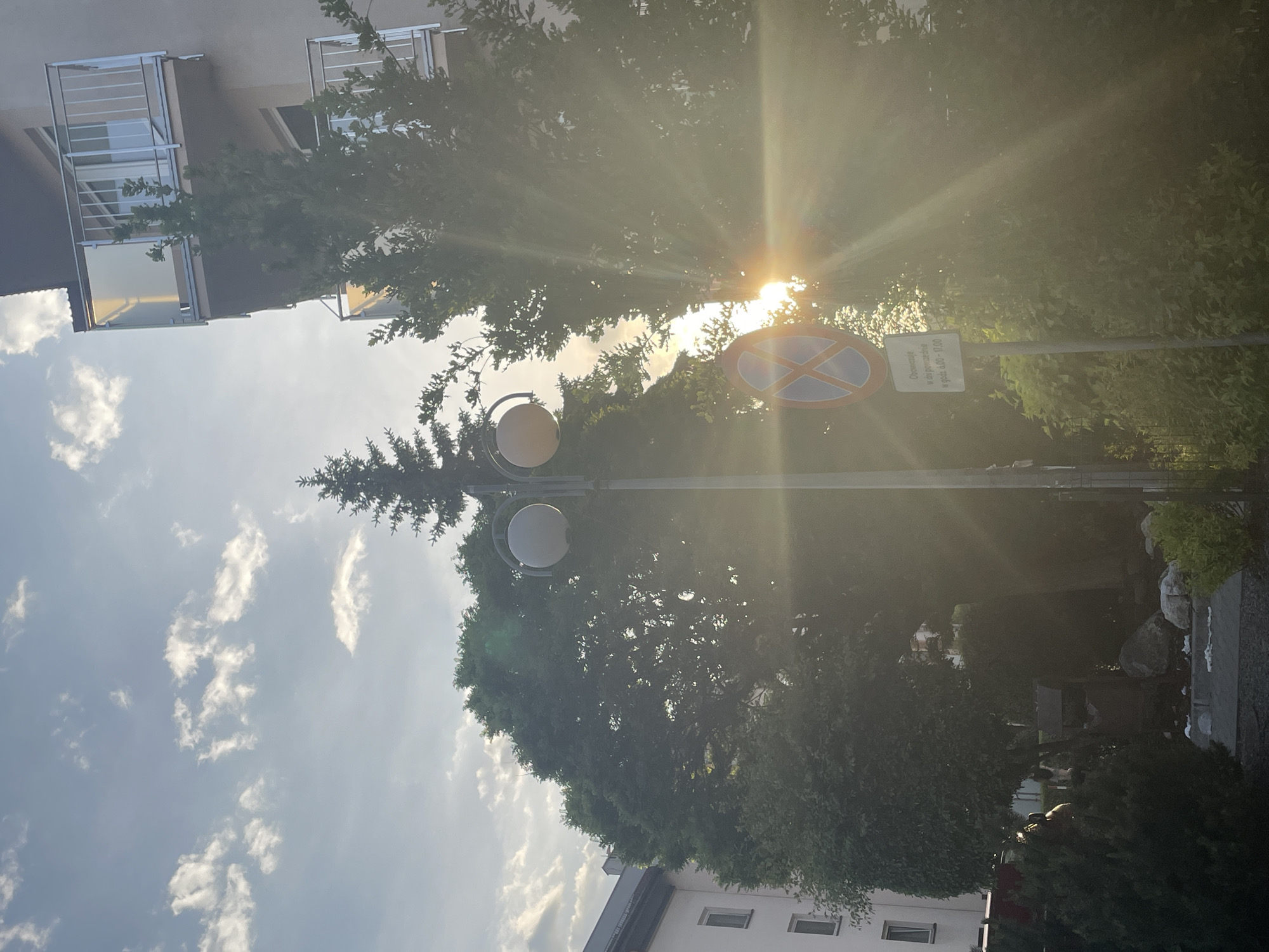 The image shows an outdoor scene with a building on the left side, a street lamp in the center, and trees filling the background. The sun shines brightly through the trees, creating lens flares. A traffic sign is attached to the street lamp pole. The sky is visible on the left with scattered clouds. The building has windows and balconies. On the right side, there is greenery, and what appears to be a stone structure in the background.