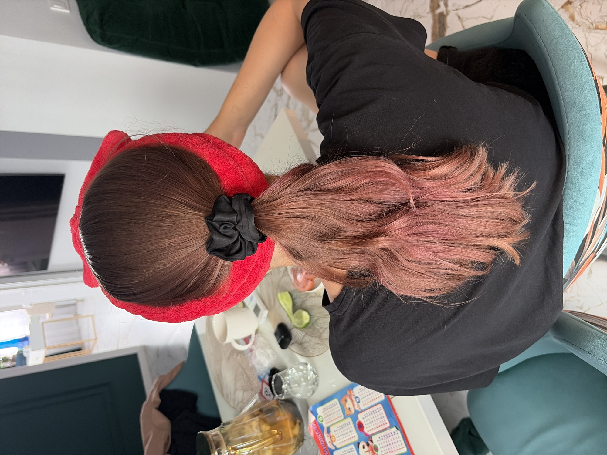 A person with long brown hair, with pinkish tips, is seated facing away from the camera, wearing a black t-shirt and a red headband that resembles cat ears. The hair is tied in a ponytail with a black scrunchie. On the table in front of the person, there is a white mug, a small clear glass, a lime wedge, and a decorative tray. A jar with a yellow liquid is also on the table. A colorful paper with cartoon characters is visible on the left side of the table. The scene appears to be indoors, possibly in a living room or dining area, with a white and marbled floor. The background shows a glimpse of furniture and a doorway. There is no information in the image to suggest the location is Moscow, Russia.