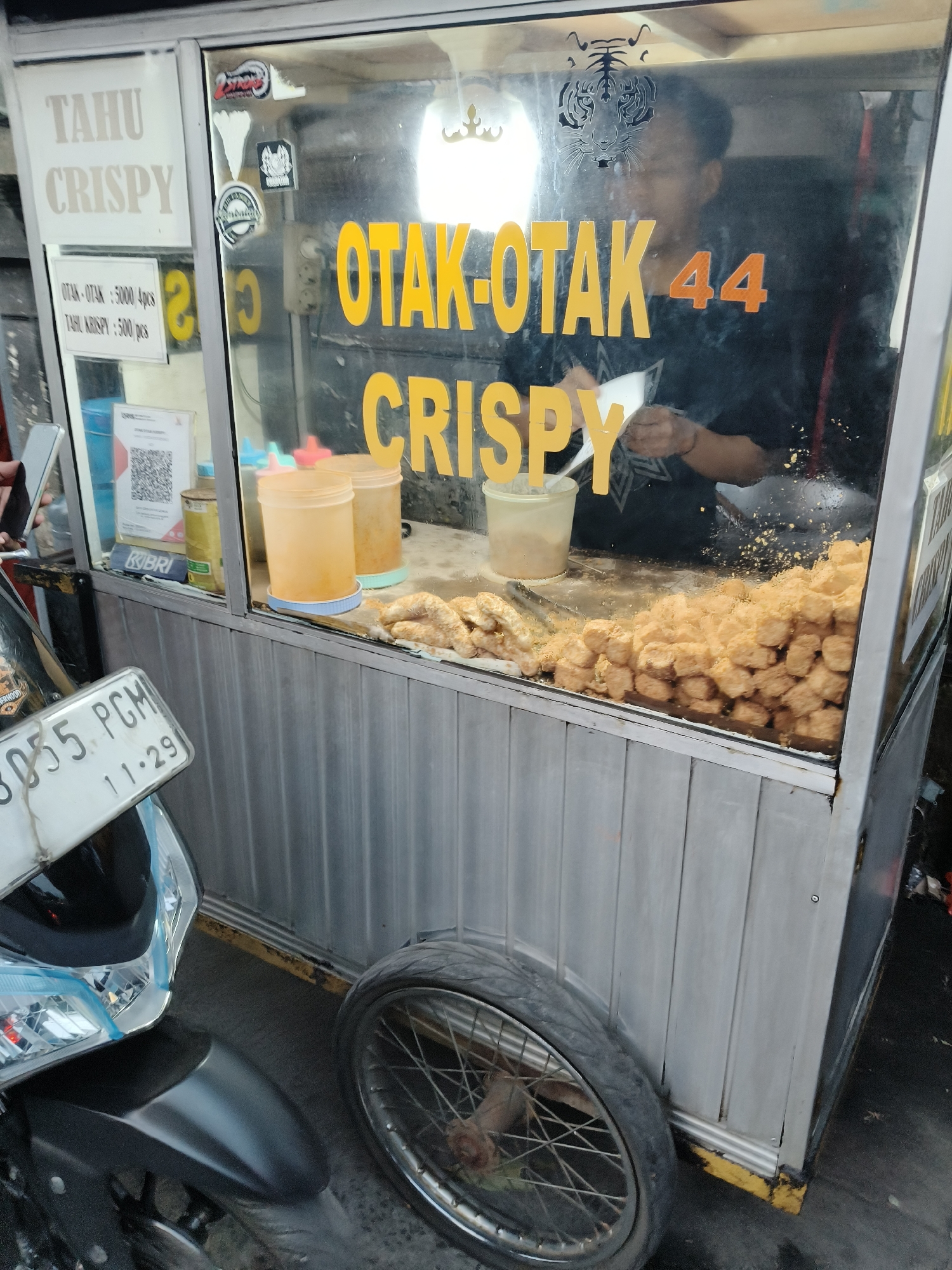 A mobile food cart selling "OTAK-OTAK CRISPY" and "TAHU CRISPY" is parked on a street. The cart features a glass display case filled with fried tofu pieces and various condiments. A man is visible behind the counter, preparing food. A motorcycle with a license plate reading "B 3055 PCM 11-29" is parked next to the cart, partially obstructing the view of a wheel. The background suggests a typical street scene in South Tangerang, Indonesia.