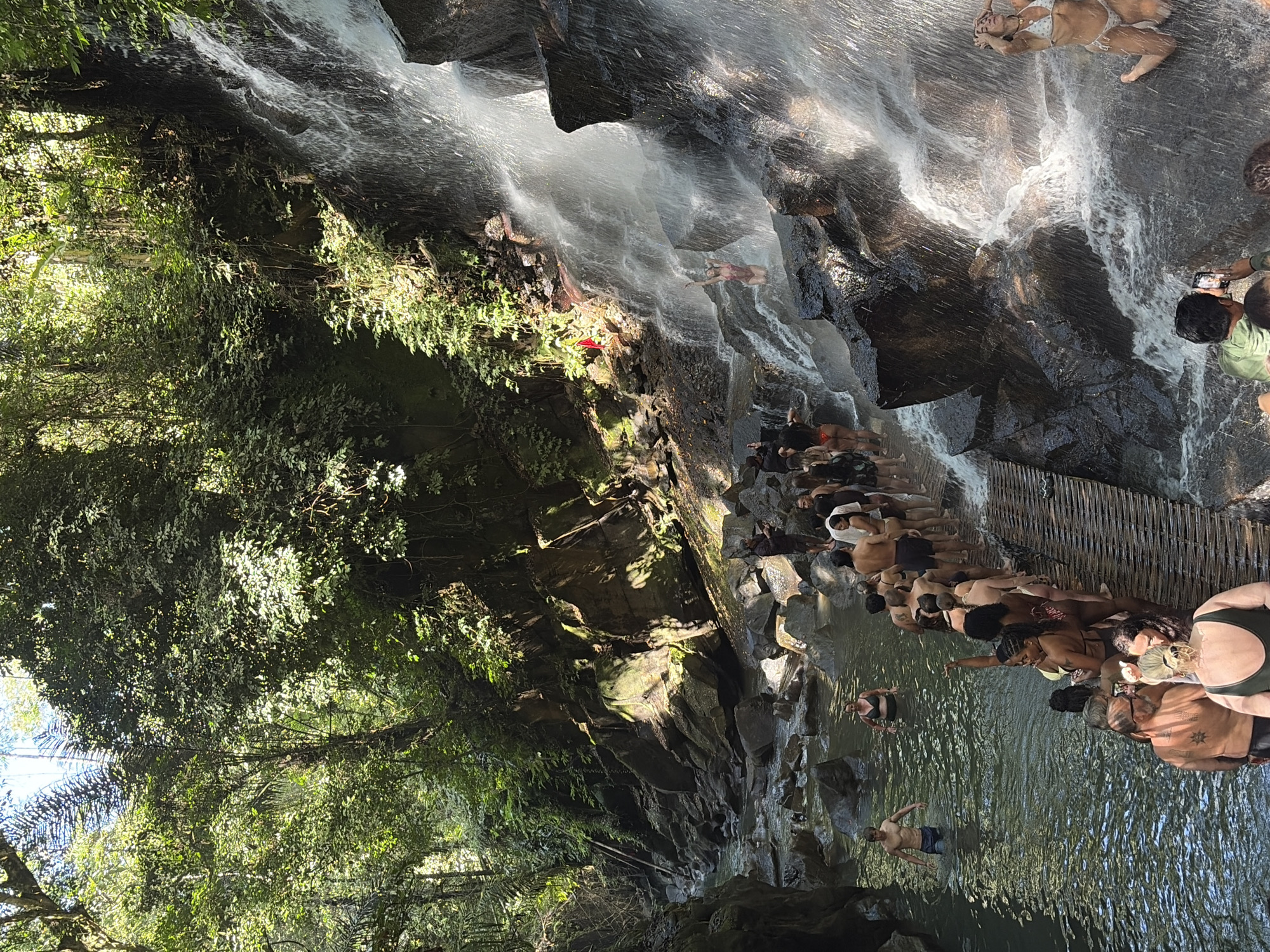 The image depicts a crowded natural setting with a cascading waterfall and a river, likely a tourist attraction in Gianyar, Indonesia. A wooden walkway follows the edge of the river, where a long line of people, predominantly in swimwear, are waiting or walking. Some individuals are swimming in the river. The waterfall features numerous people interacting with the water, some standing directly under the falls, others posing for photos. The environment is lush and green, with dense foliage and large rocks surrounding the waterfall and river. Sunlight filters through the trees, creating dappled light and shadows. The overall scene conveys a sense of activity and enjoyment in a tropical, natural landscape.