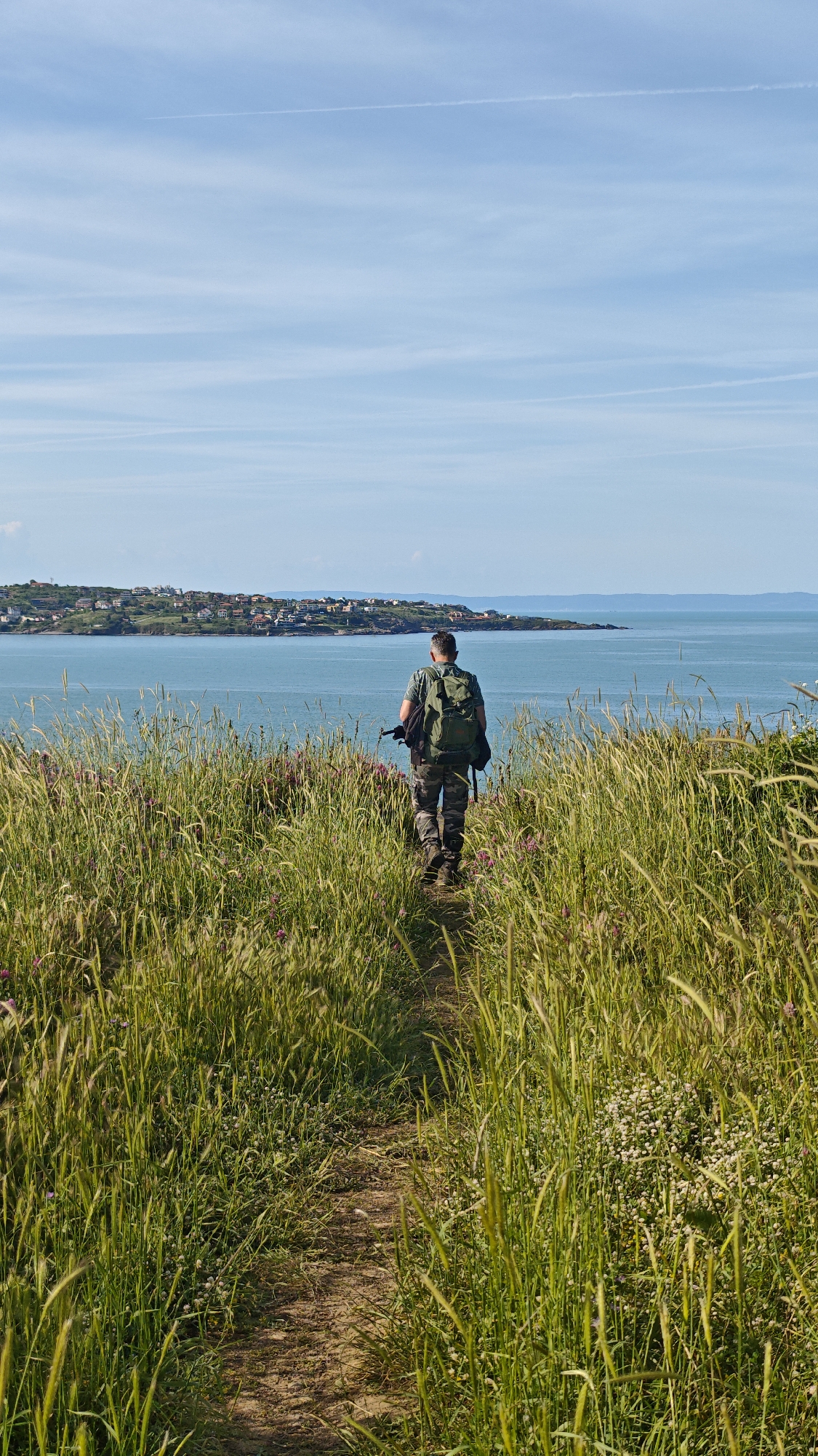 A medium, full shot captures a man from behind as he walks along a narrow dirt path through tall, green grass and wildflowers toward a town along the coast. The man wears a short-sleeved green shirt, camouflage pants, and hiking boots, and a green backpack is strapped to his back. He is fair-skinned with short, graying hair. The path is surrounded by dense, sunlit grasses and small white and purple flowers. Beyond the grass is a blue sea, and across the water, a town with numerous red-roofed buildings is visible. The sky is light blue with a few wispy clouds. The scene is likely located in or near Sozopol, Bulgaria, due to the coastal town architecture and landscape features common to the area.