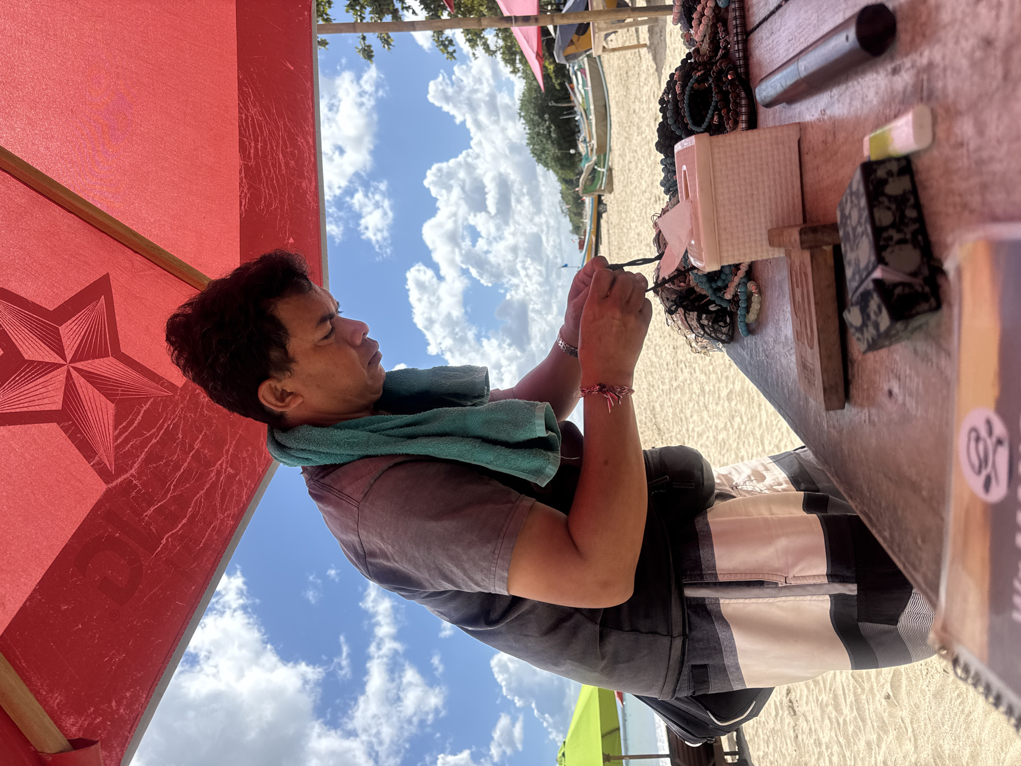 A man is crafting beaded bracelets under a red umbrella on a sandy beach. He wears a gray t-shirt, black shorts with white and gray stripes, and a turquoise towel draped around his neck. In front of him is a table with various items including a tissue box, several piles of beaded bracelets, a wooden block, a metal tube, and a small box that appears to be a cigarette case. The background shows the ocean, traditional Balinese boats, and green trees, indicating the location is Kuta Selatan, Indonesia. The sky is blue with scattered white clouds.