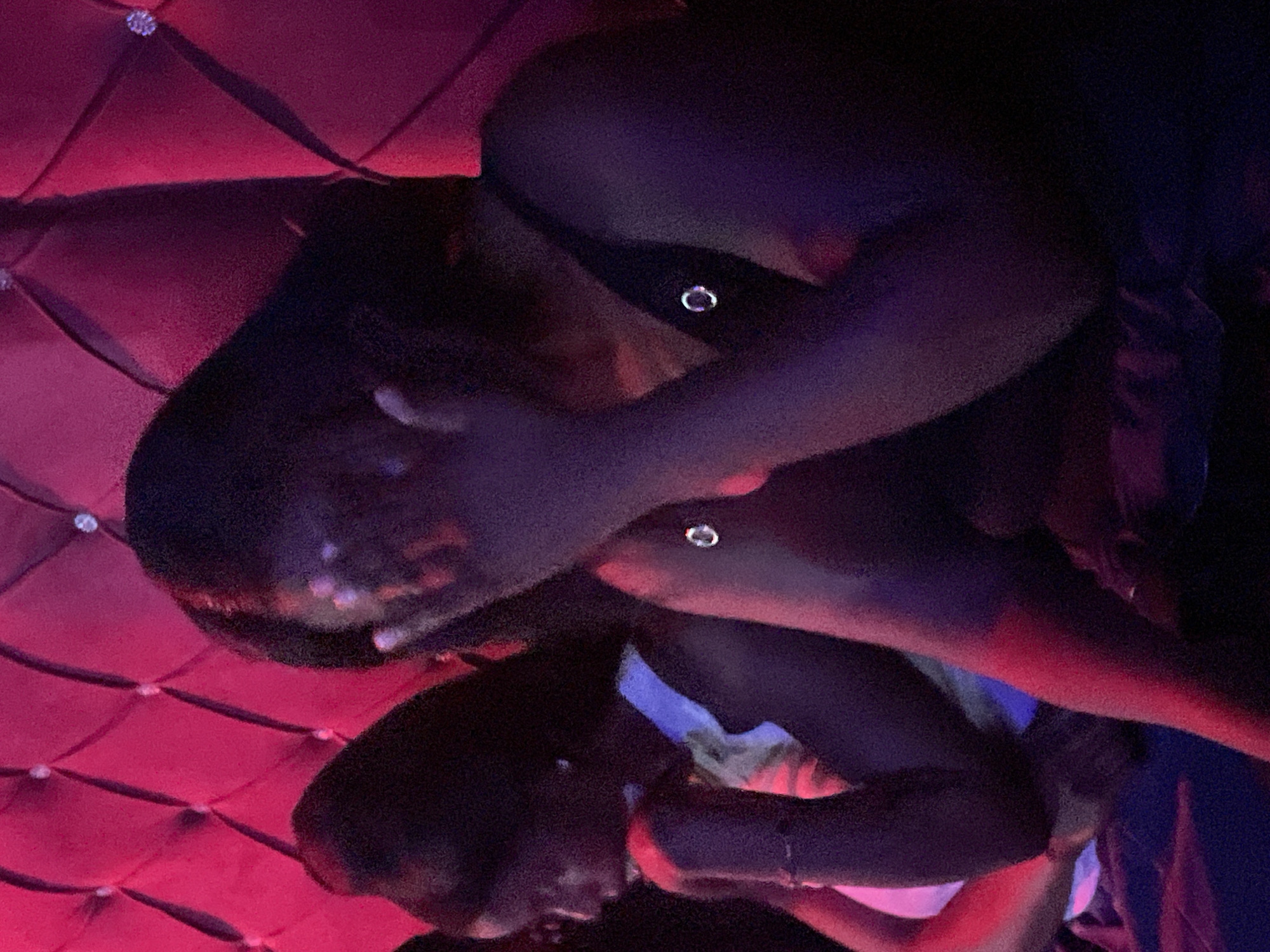 The image depicts two individuals seated against a vibrant pink tufted backdrop, likely a booth or seating area in an establishment within Durumi, Nigeria. The lighting is dim and colored, casting a purplish hue over the scene. The person in the foreground, on the right, is partially obscured by their hand covering their face. They are wearing a dark tank top with a circular metal embellishment. To the left, another individual is visible, looking downwards and resting their chin on their hand. They are wearing a white top with yellow accents and a bracelet on their wrist. The environment appears to be a dimly lit social setting, possibly a club or lounge, characterized by the plush, diamond-tufted pink wall.