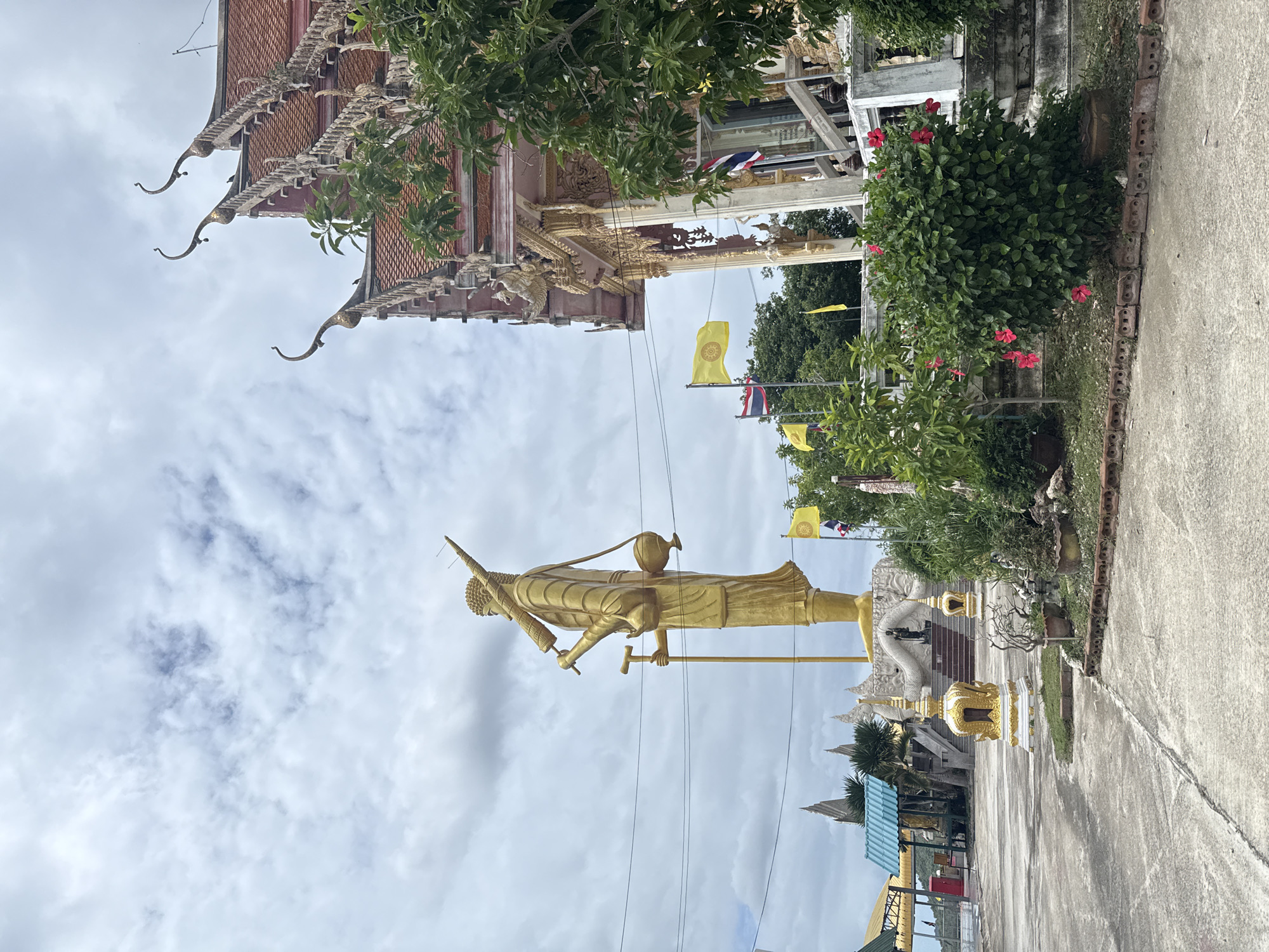 The image depicts a scene outside a Buddhist temple in Hua Hin City Municipality, Thailand. Dominating the left side of the frame is a large, golden statue of a standing figure, possibly a monk, carrying a staff and a bowl. Behind the statue, to the right, is a building with traditional Thai temple architecture, featuring ornate roofs and decorative carvings. Several yellow flags, one with a Buddhist emblem, and Thai flags are visible, some flying from poles and others attached to the building. Green foliage, including a flowering bush with red hibiscus, surrounds the temple building and the base of the statue. The ground is paved with concrete, and power lines are visible crossing the sky. The sky is overcast with clouds. The overall impression is one of religious significance and cultural representation within a tropical setting.