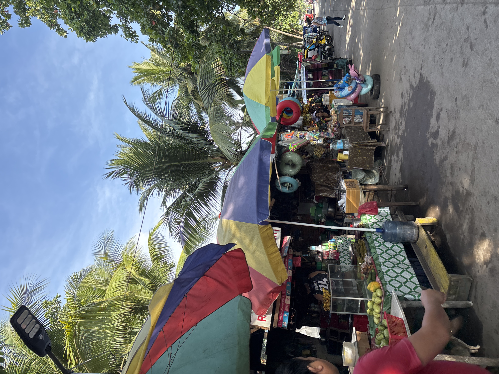 This is a daytime, eye-level outdoor street scene in a tropical location, possibly Davao City, Philippines. The foreground shows a person in a red shirt looking towards a market stall that is covered by a large, colorful umbrella. The stall displays various items, including fruits like green mangoes and bananas, bottled drinks, and other goods. Behind the stall, more market stalls and palm trees are visible, creating a lush, tropical atmosphere. The street is paved and appears to be a public area with a few motorbikes and a person walking in the distance. The overall scene depicts a vibrant street market in a warm climate.