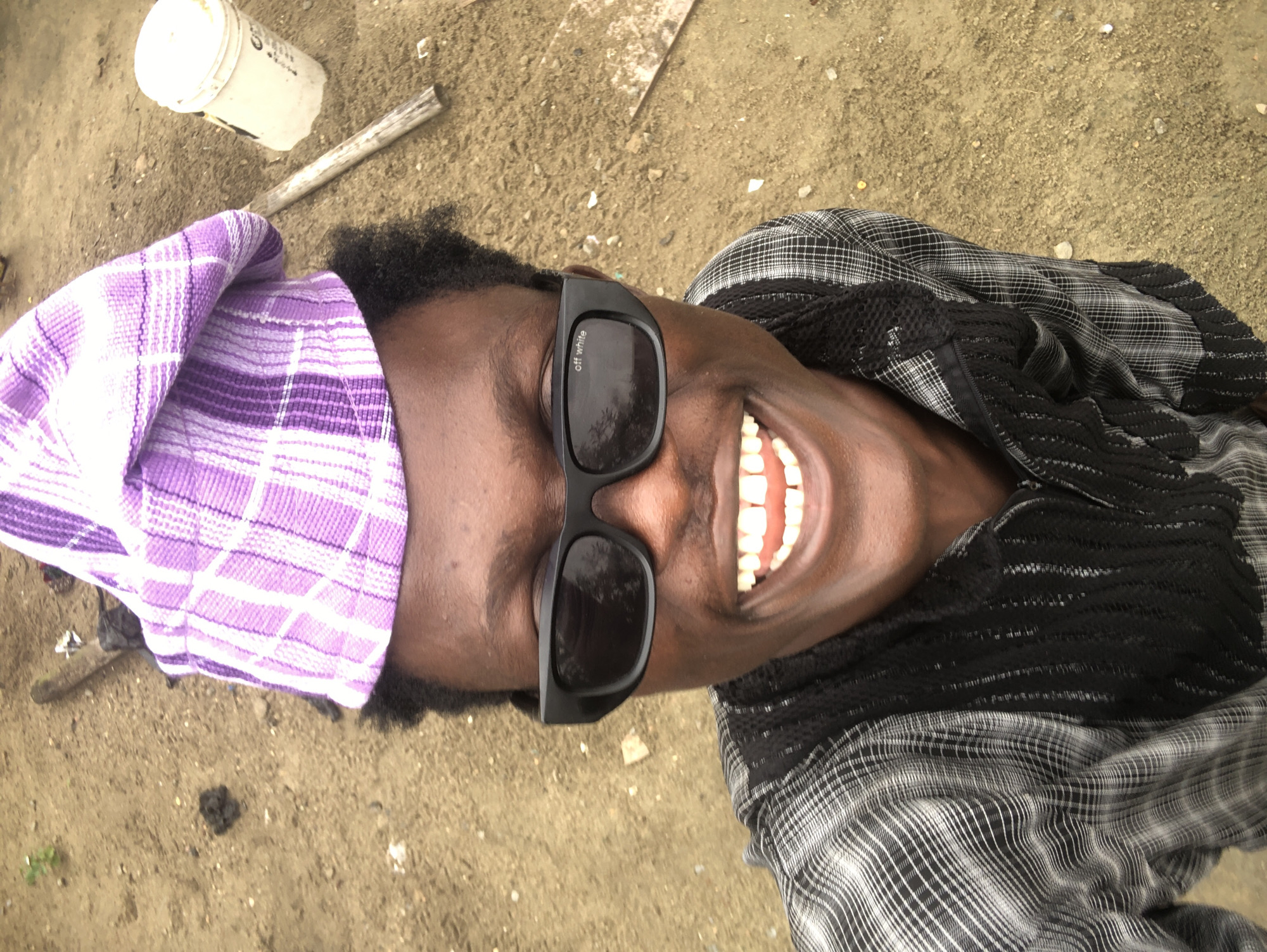 A young Black male wearing a purple and white checkered cap, black sunglasses, and a black and white checkered shirt with a black lace trim is smiling broadly at the camera. The man is outdoors, with a sandy ground and a few scattered rocks and debris visible. In the background, a white bucket and a wooden stick are also present. The overall setting appears to be an unpaved area, possibly a construction site or a rural road in Ado, Nigeria, given the dry, dusty earth and the simple, utilitarian objects. The lighting suggests it is daytime.