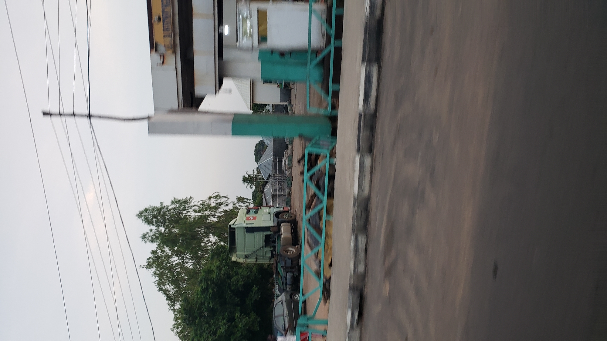 This image captures a scene in Jalingo, Nigeria. In the foreground, a blurred street surface suggests movement. A light-colored curb with black stripes runs along the edge of the road. Beyond the curb, a turquoise metal barrier with a geometric pattern is visible.
In the middle ground, a light green truck cab is parked on a dirt or gravel lot. The truck has a distinctive red emblem on its side. To the left of the truck, a dark-colored car is parked. Behind the truck and car, several structures with corrugated metal roofs are visible, resembling temporary shelters or market stalls. Trees, some with lush green foliage, are present in the background.
To the right, a structure with white and green columns stands prominently. This appears to be part of a building, possibly a filling station or a commercial establishment. A white wall with a white rectangular panel is also visible. Overhead, several power lines crisscross the sky, which is overcast. The overall impression is of a bustling area with a mix of vehicles, buildings, and vegetation.
