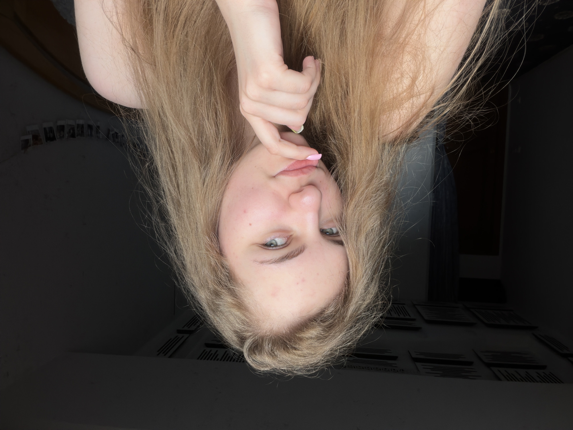 A young woman with long, blonde, disheveled hair is pictured from the chest up, looking directly at the camera. She has light skin with some blemishes on her forehead and cheeks, and blue-green eyes. Her right hand is raised, with her index finger gently touching her chin. Her fingernails are painted pink. She is wearing a light-colored top, possibly off-the-shoulder, though only her left shoulder is visible. The background is dimly lit, with dark walls and a ceiling adorned with what appear to be acoustic panels or decorative tiles. A string of small framed photos hangs on the wall to the right. The setting is likely a bedroom or personal living space, not identifiable as being in the city of Dnipro, Ukraine, based solely on the visual information.