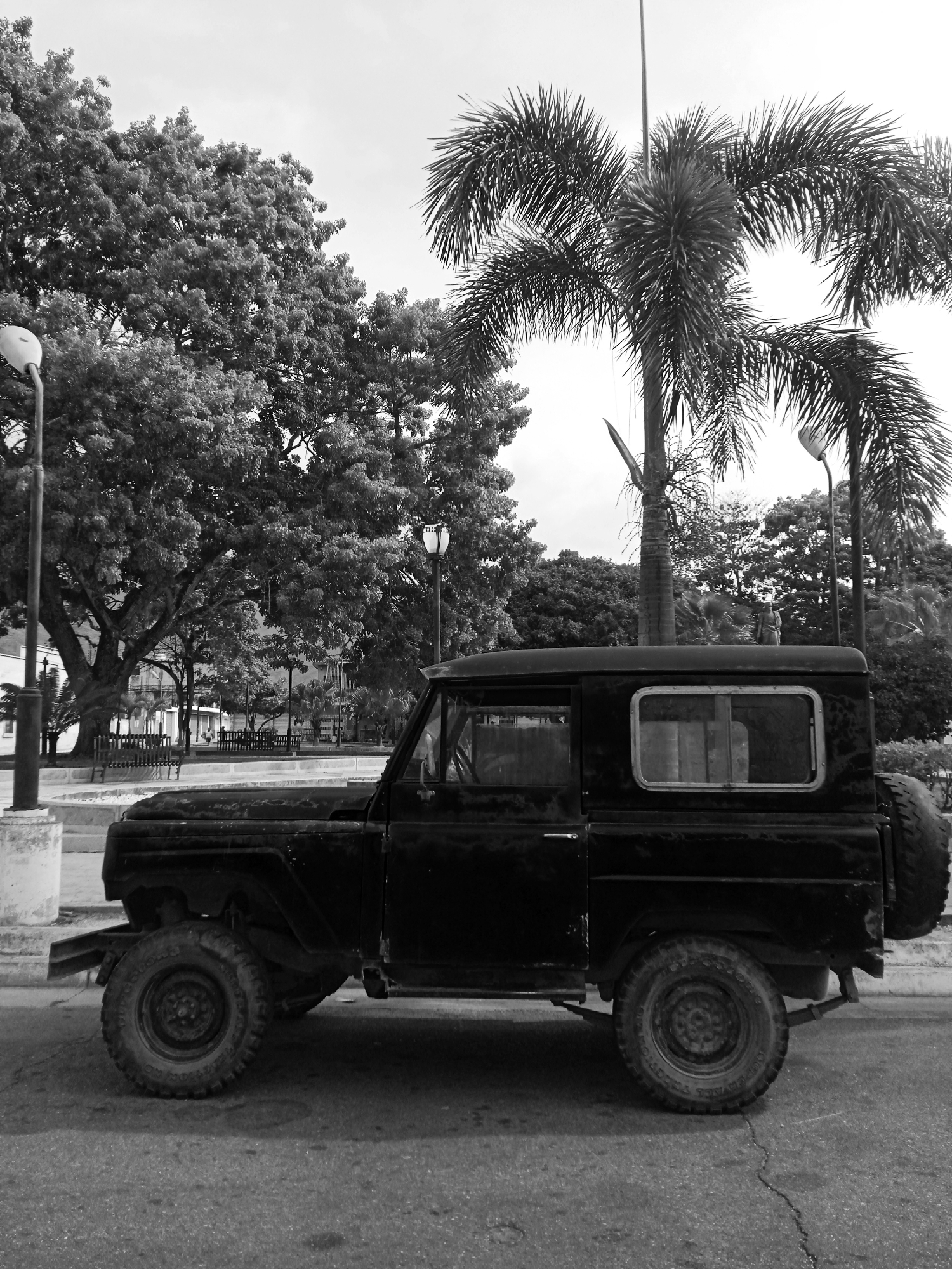 The image is a black and white photograph depicting an old, black off-road vehicle parked on the side of a cracked paved road. The vehicle has large tires and a spare tire mounted on the back. In the background, there is a park-like area with large trees, including a palm tree. There are lamp posts visible in the background and a bench in the park. The location is Montalbán, Venezuela.