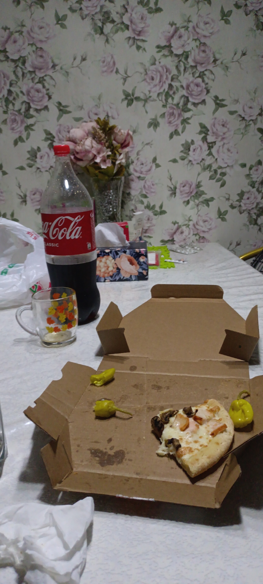 The image depicts a tabletop scene with food and drink, set against a floral wallpaper background. In the foreground, a partially eaten slice of pizza sits in a cardboard pizza box. Two yellow pickled peppers are placed near the pizza slice. To the left of the box, a glass mug with a fall leaf pattern is visible, and next to it, a large bottle of Coca-Cola Classic. A white paper bag and a box of tissues with a floral design are also present on the table. The scene is domestic and appears to be an informal meal setting. There are no discernible landmarks or details to suggest a specific location within Fergana City, Uzbekistan.